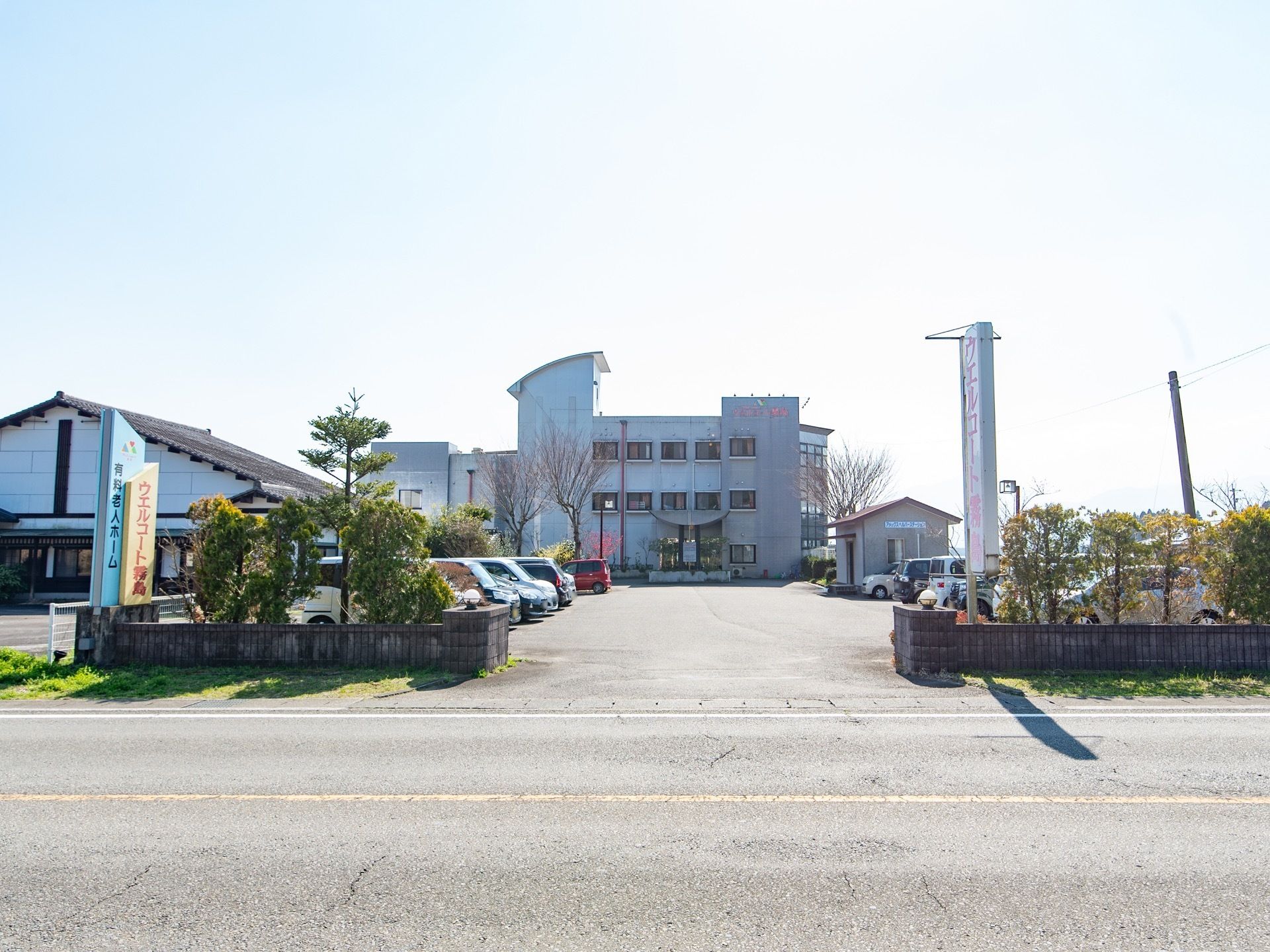 片側１車線の道路の向こう側に３階建ての建物が建っている。建物の前にたくさんの車が停められた駐車スペースがある。