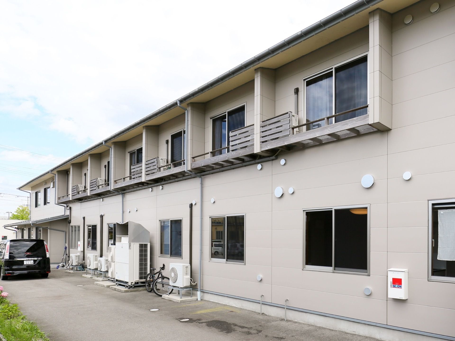白を基調とした２階建ての建物である。外壁に沿って複数のエアコンの室外機が設置されており、自転車と車が停められている。