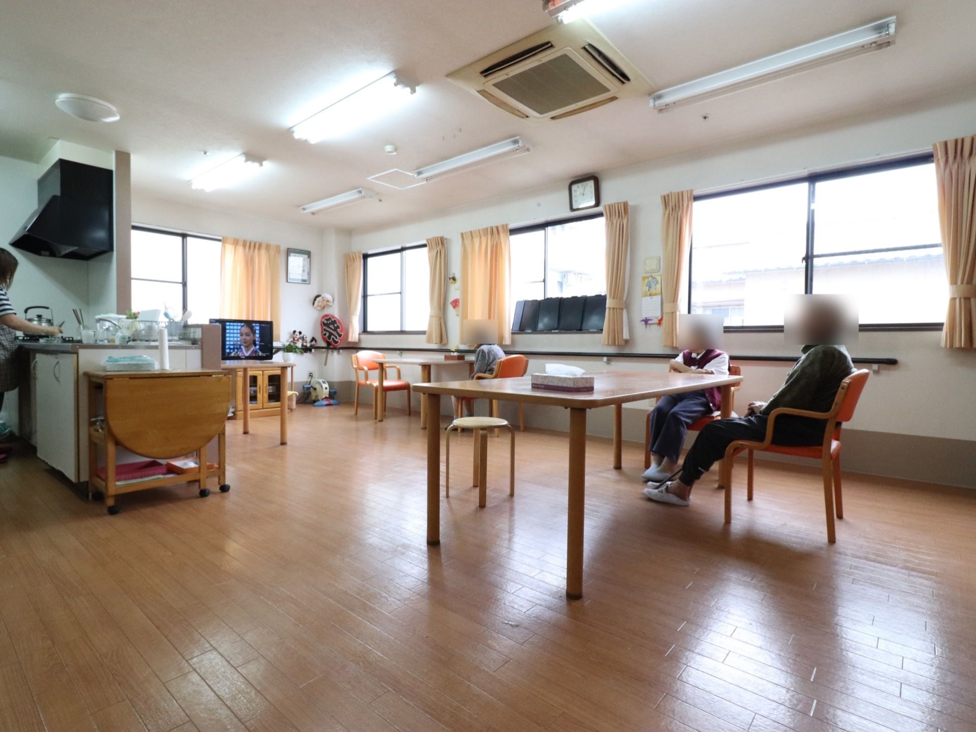 腰窓がある明るい空間に、ダイニングテーブルと椅子が備わっている。テレビがあり利用者様が鑑賞している。対面キッチンと隣接している。