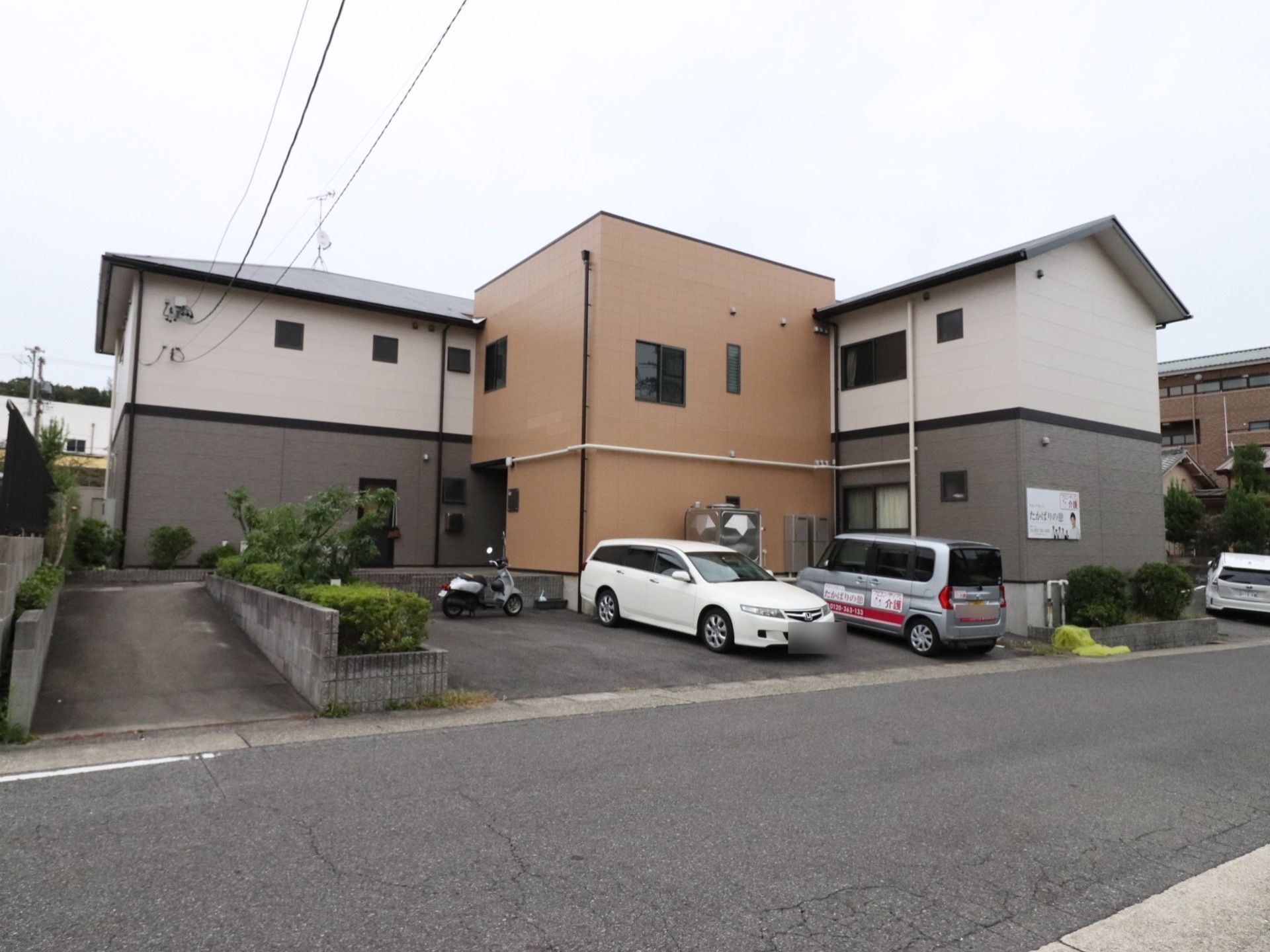 施設の外観である。2階部分が白色で1階部分がグレーの外壁で、エントランスがある建物は全体が茶色の外壁である。