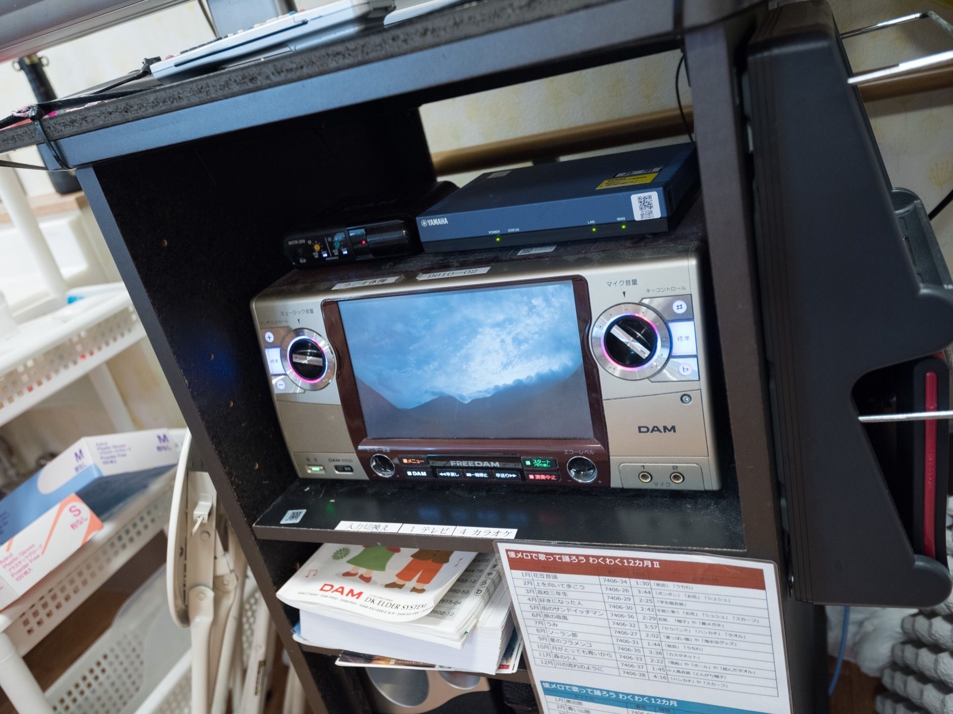 カラオケマシーンは、丸いハンドルを回してマイクの音量などを調節する。黒い棚に収めてあり、曲の番号が記載してある本も積んである。