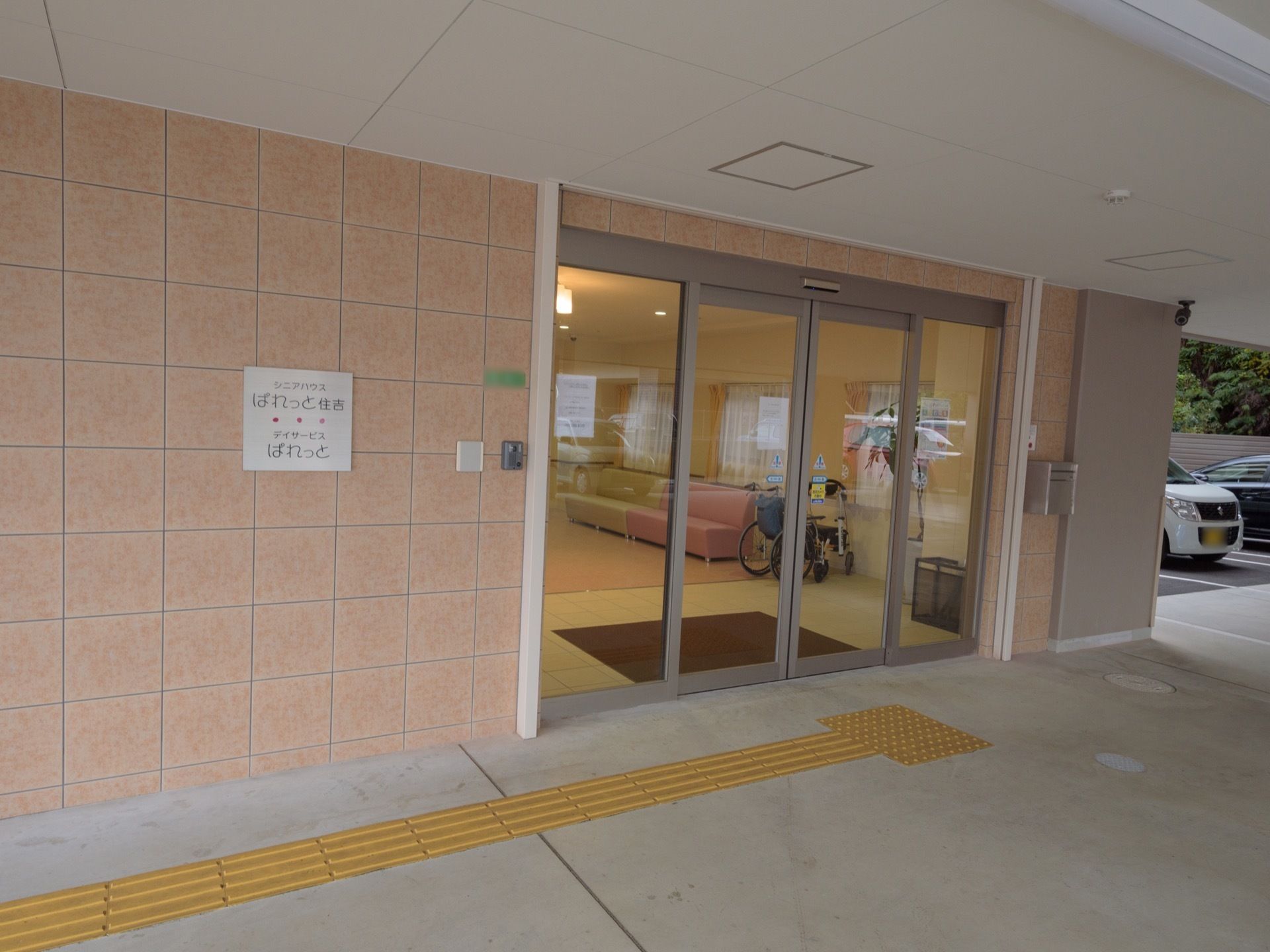 施設の入口は大きく開くガラスの自動ドア。入口前は大きな屋根が付いている。外壁に施設名が書かれた表札が付いている。