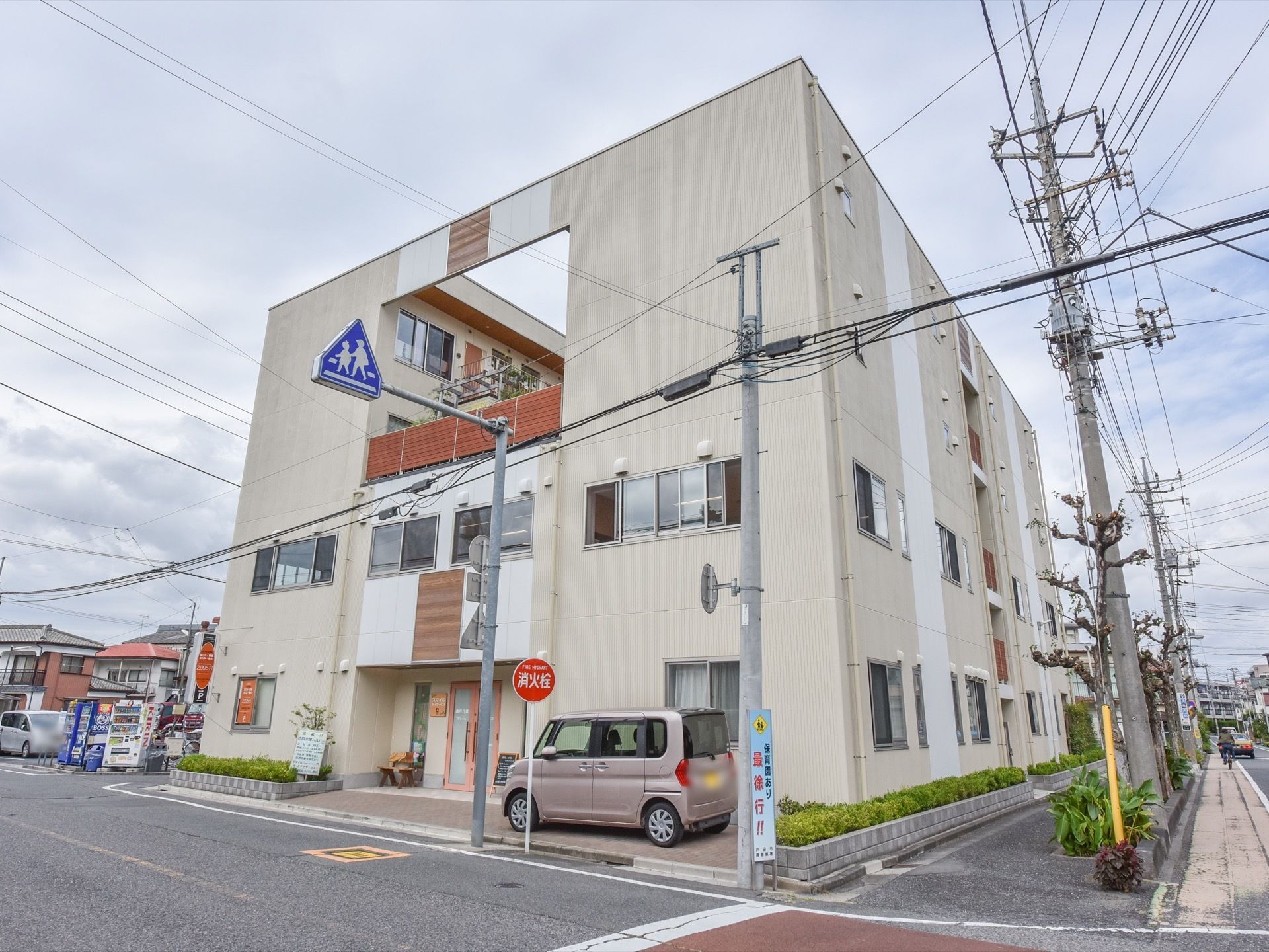 施設は道路に面して建てられている。道路には電柱がたくさん設置されている。建物の入り口扉の横には車が止められている。