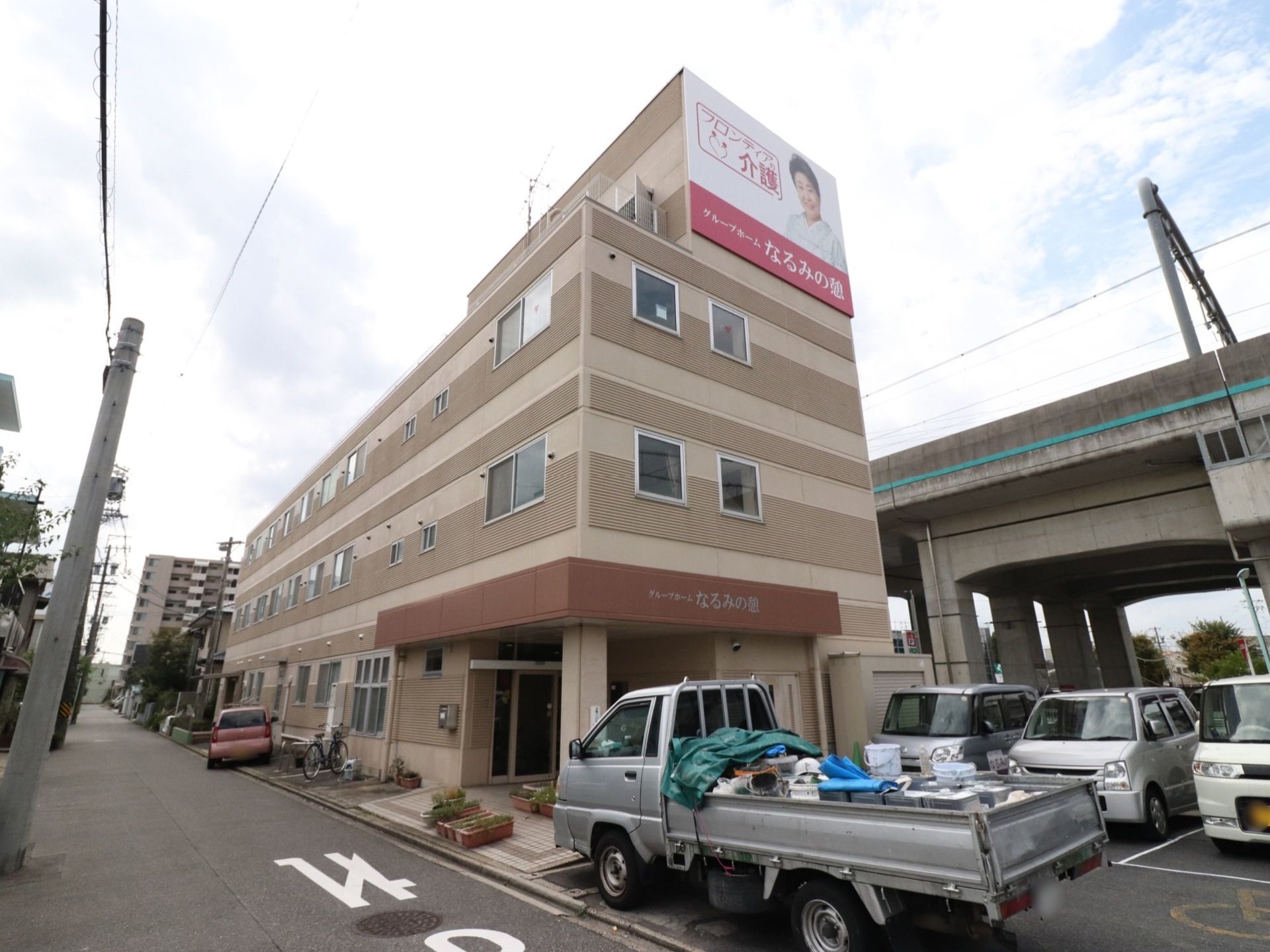 落ち着いた色の３階建ての建物。入口前には広いスペースがあり、屋根が付いている。建物の横には数台停められる駐車場がある。