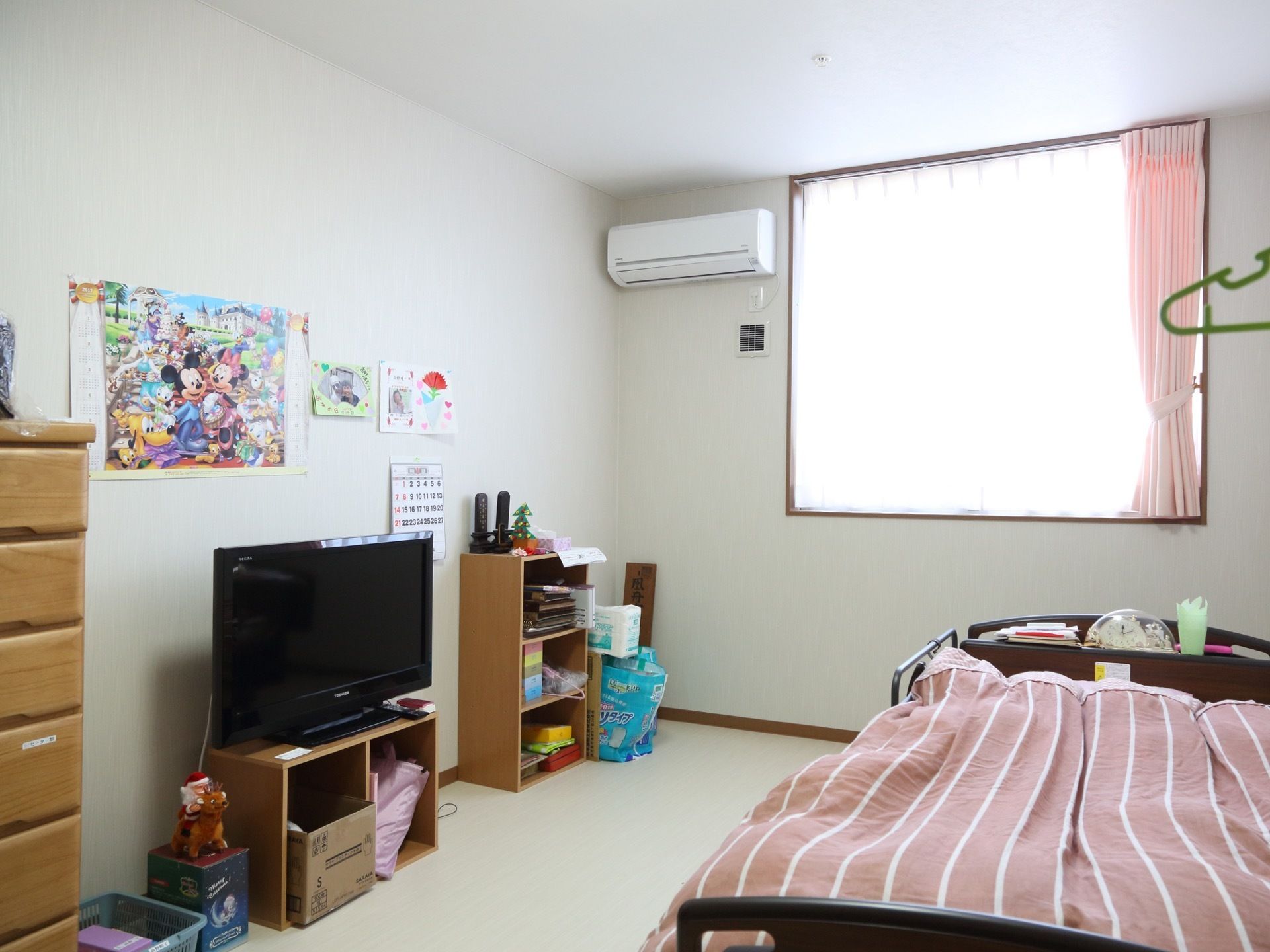 居室には、エアコンが完備されている。介護ベッドやタンス、カラーボックスが置かれている。明るい日差しが差し込む窓がある。