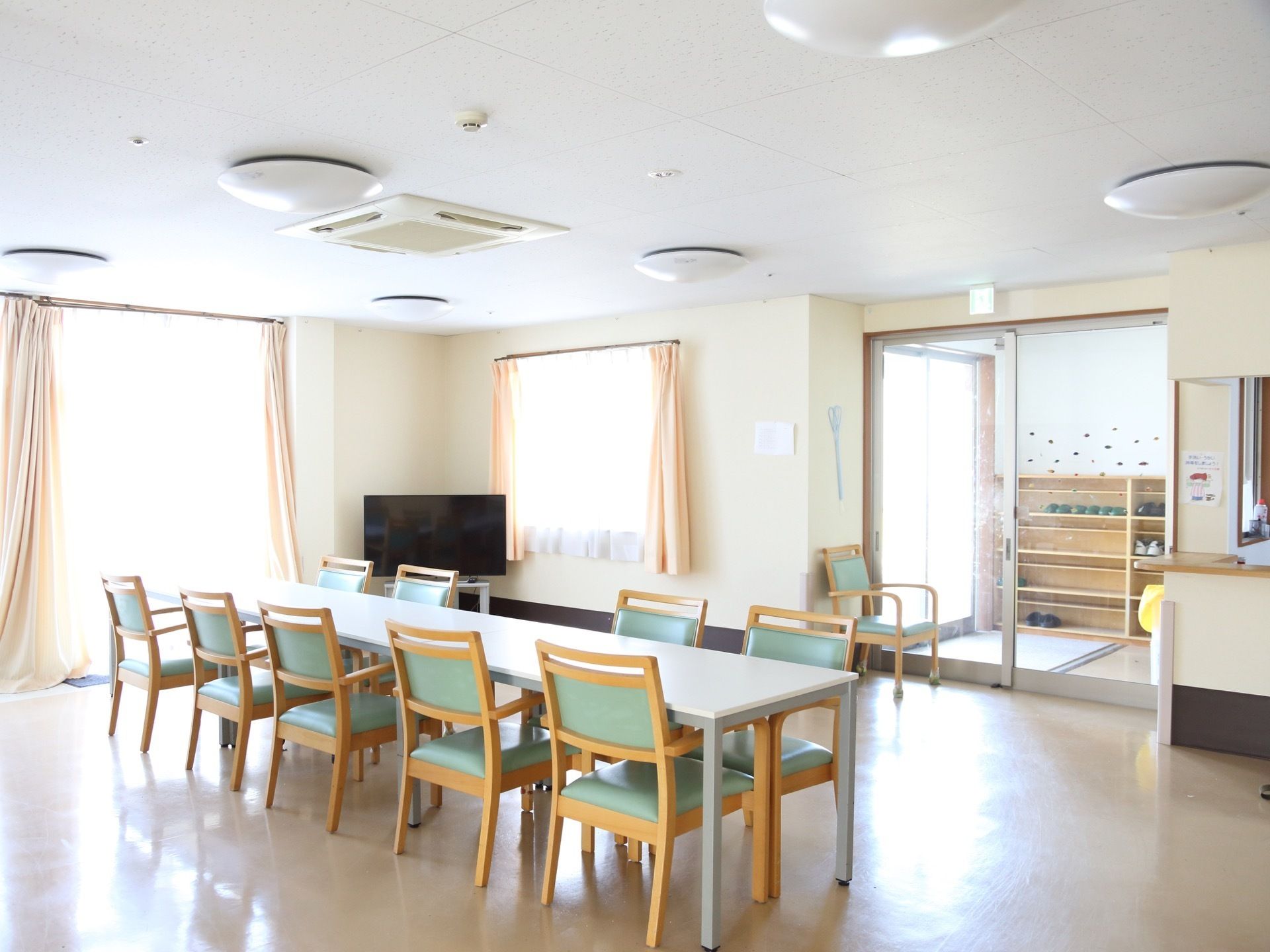 広々とした食堂の一角。窓が2か所に設置されている。中央には長机が配置されている。部屋の角にはテレビが置かれている。