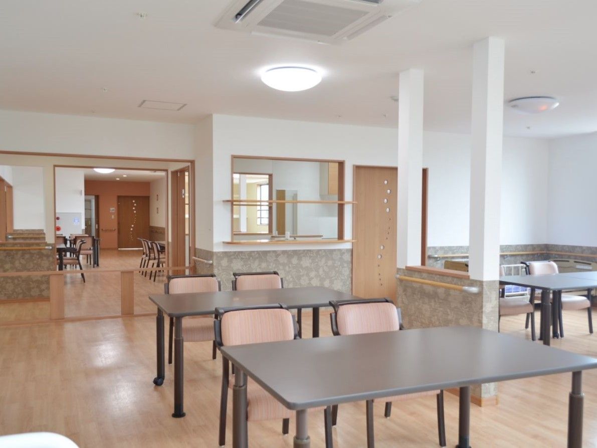 モダンなテーブルと椅子が間隔をおいて置かれている、食事や団欒が出来る空間。スタッフルームもあり、入居者様の様子を見ている。