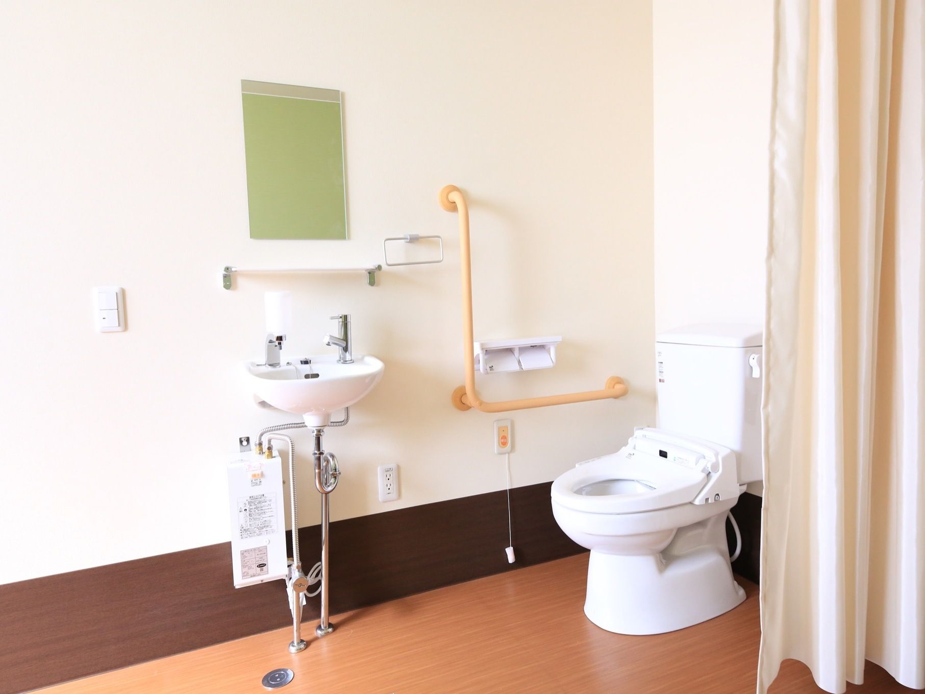タンク付きの洋式トイレの横にクリーム色のカーテンが掛けられている。壁に洗面台が設置され、鏡が取りつけられている。