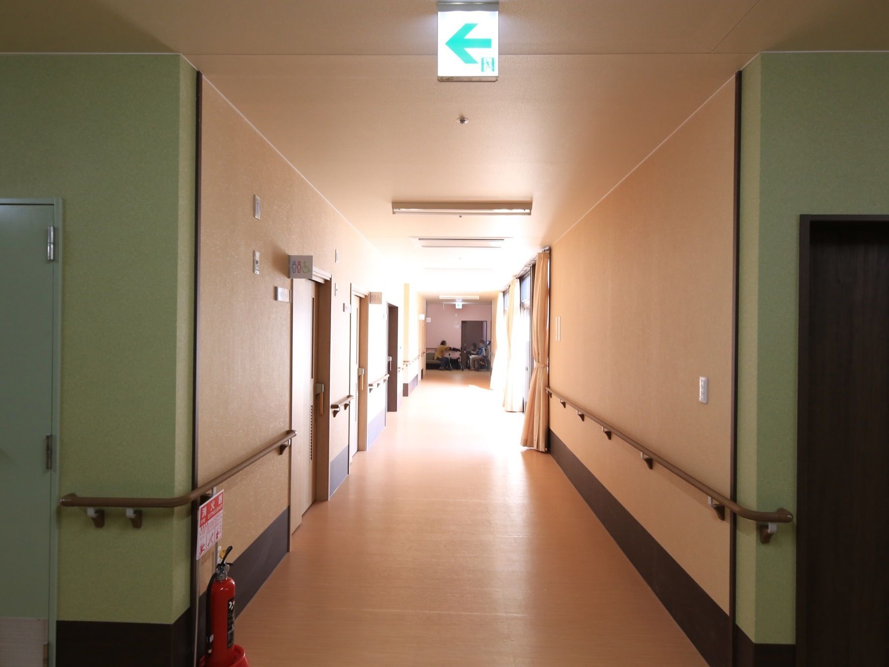 長い通路の片側に掃き出し窓があり、明るい日差しが差し込んでいる。床に消火器が置いてあり、壁に手すりが設置されている。