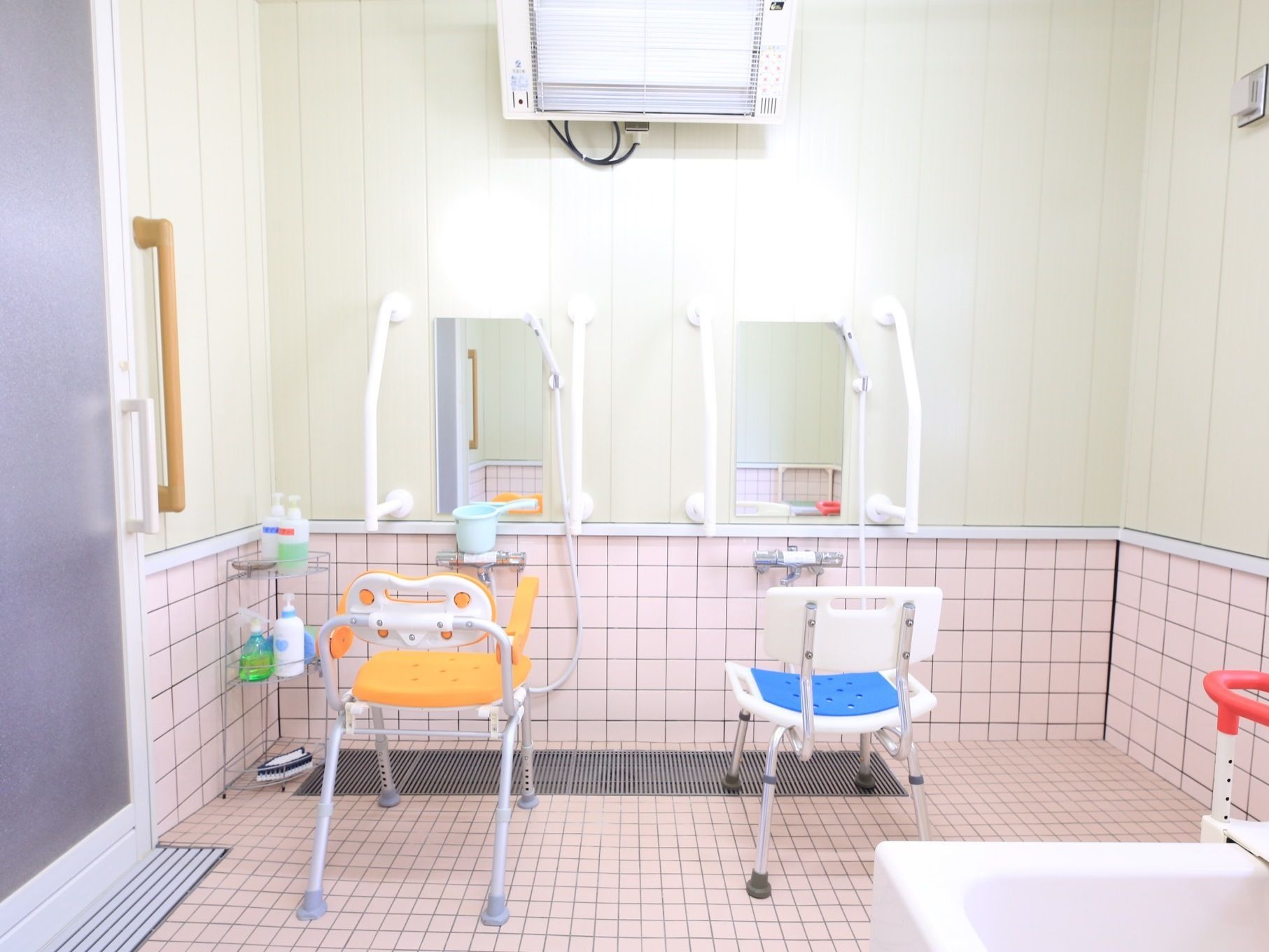 洗い場の床にピンク色の細かいタイルが貼られている。オレンジ色と青色のシャワー椅子があり、壁に鏡が設置されている。