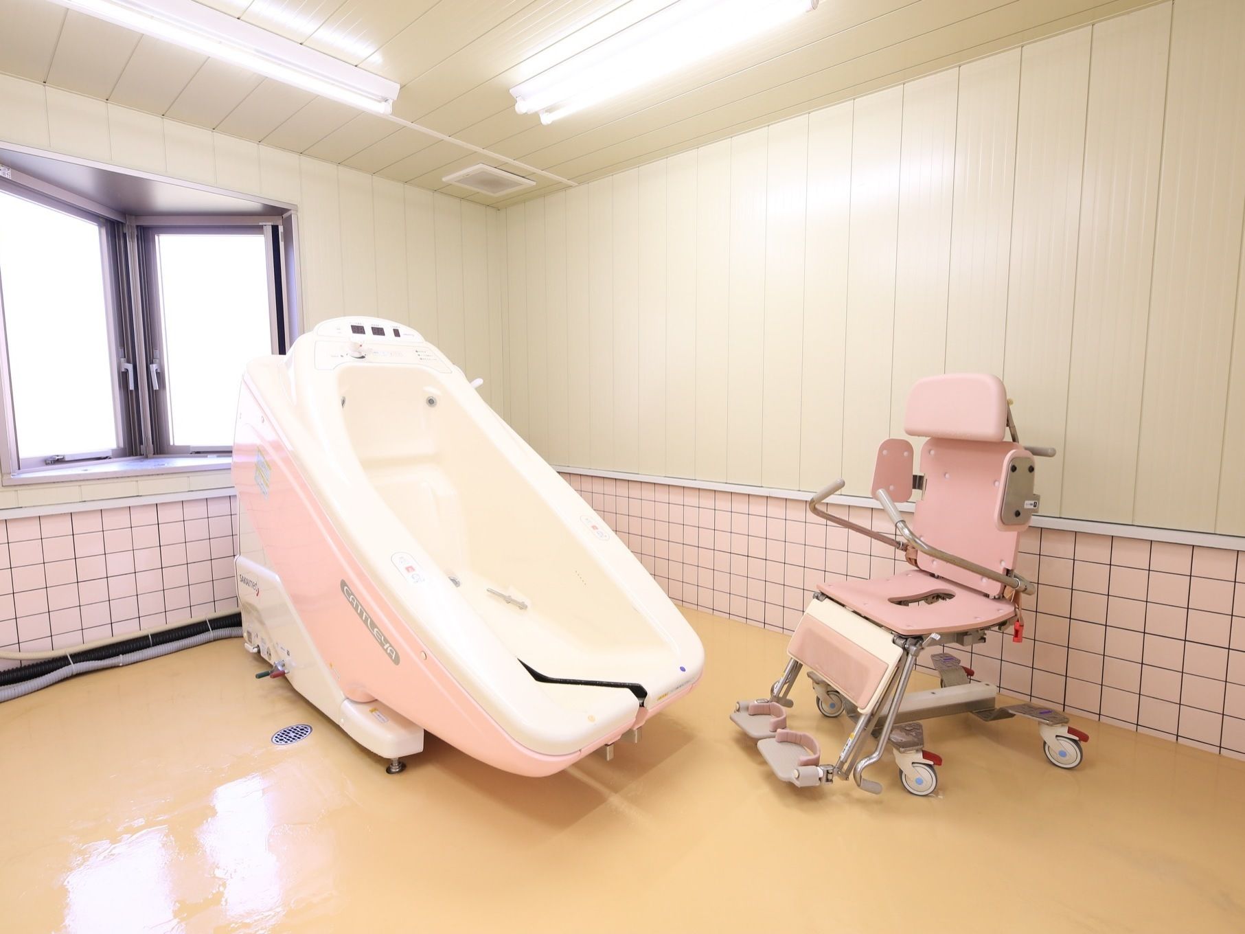 クリーム色のピカピカに磨かれた床の上に、白とピンク色の介護浴槽がある。車輪がついたピンク色の椅子が置いてある。