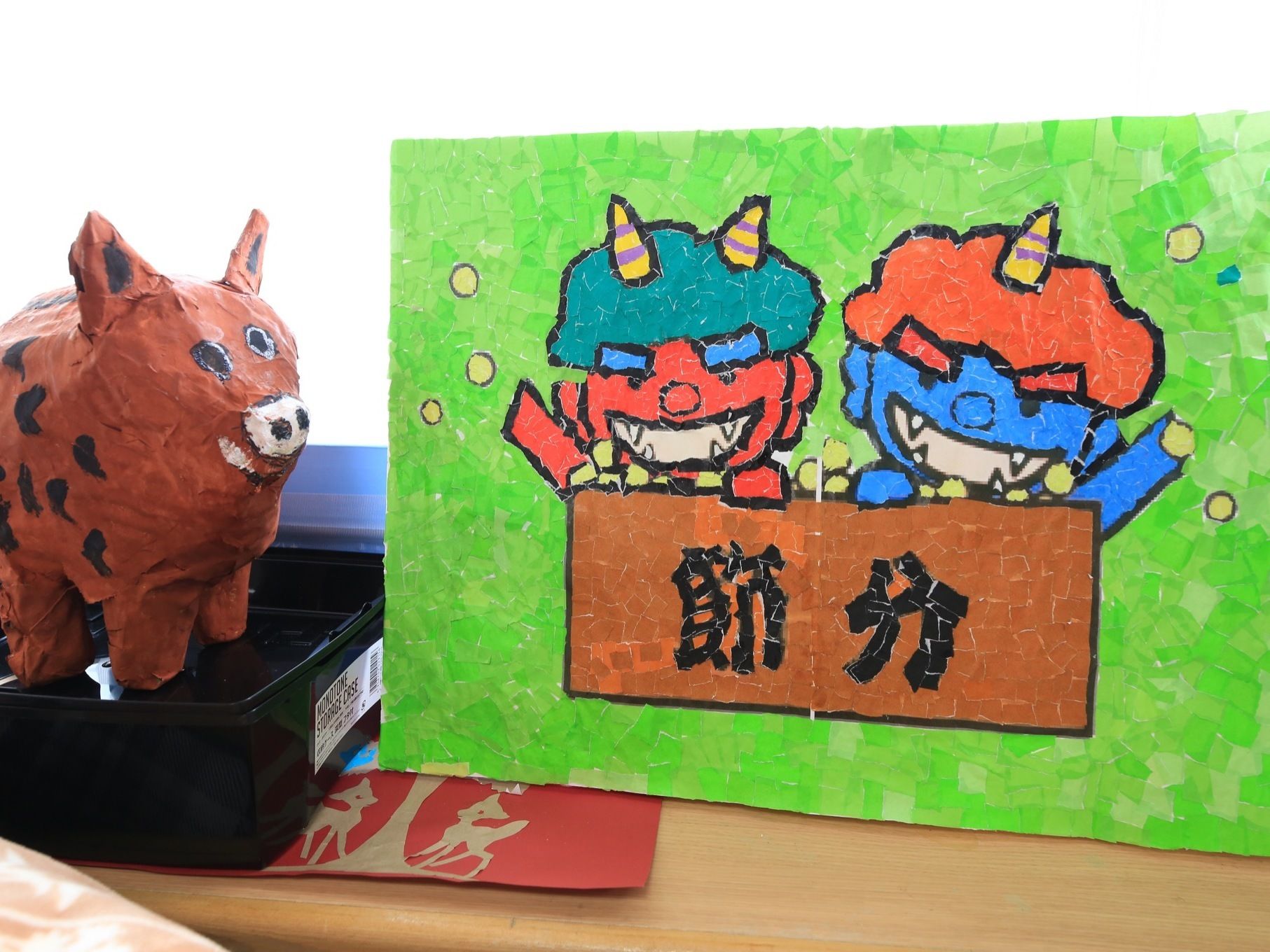 ２体の赤と青の鬼と節分の文字が切り絵で作られている。イノシシのような張り子の置物が黒い台の上に置いてある。