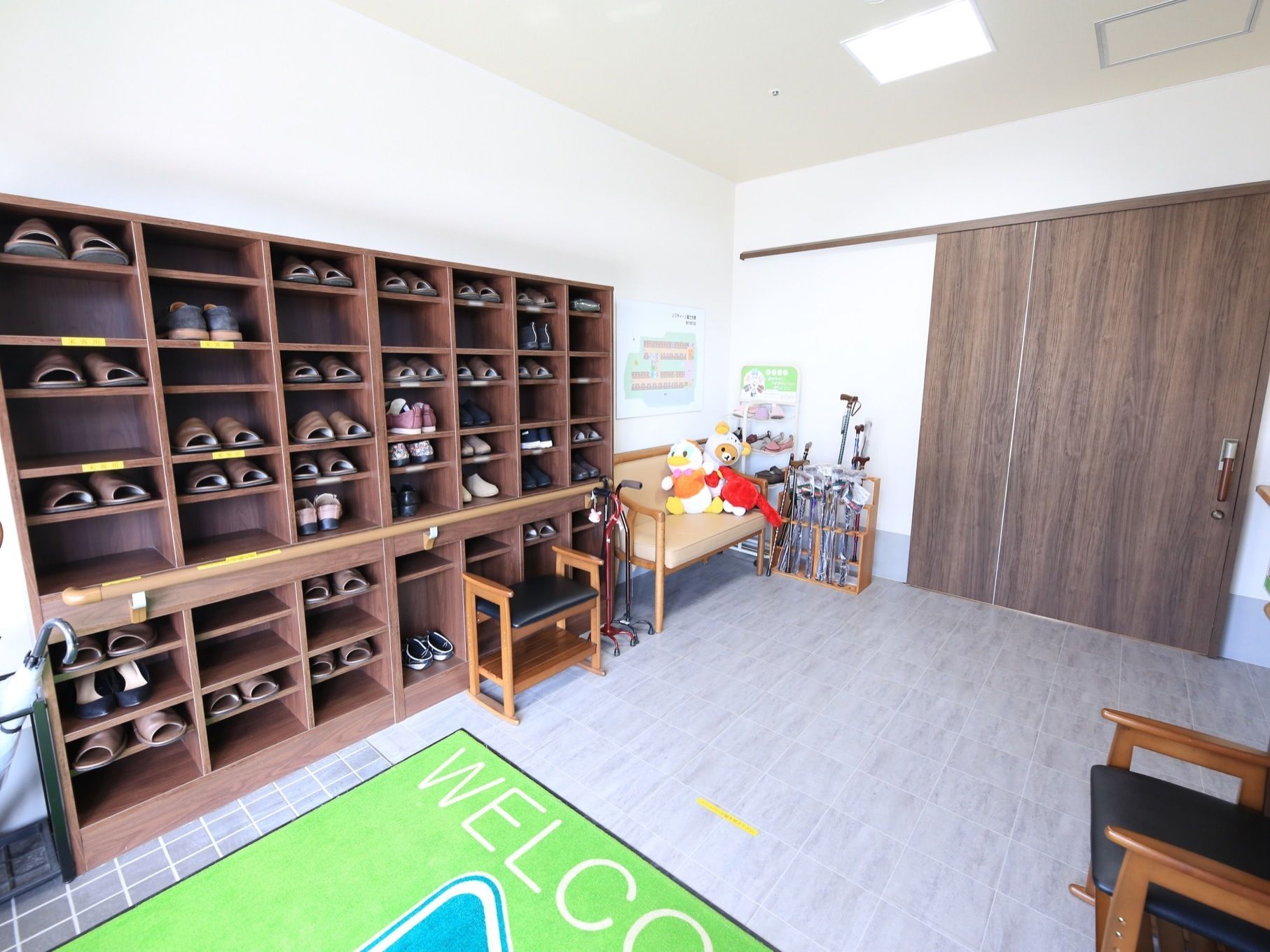 灰色の床にきみどり色の玄関マットが敷いてある。大きな靴箱にたくさんのスリッパが収納されている。椅子の上にぬいぐるみが置いてある。