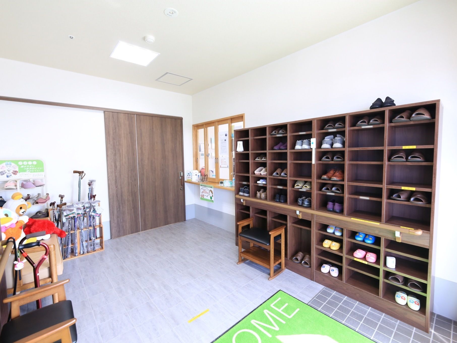 壁際に茶色い大きな靴箱が設置されている。その隣に小窓があり、窓が閉められている。茶色いスライドドアの近くにたくさんの杖が置いてある。