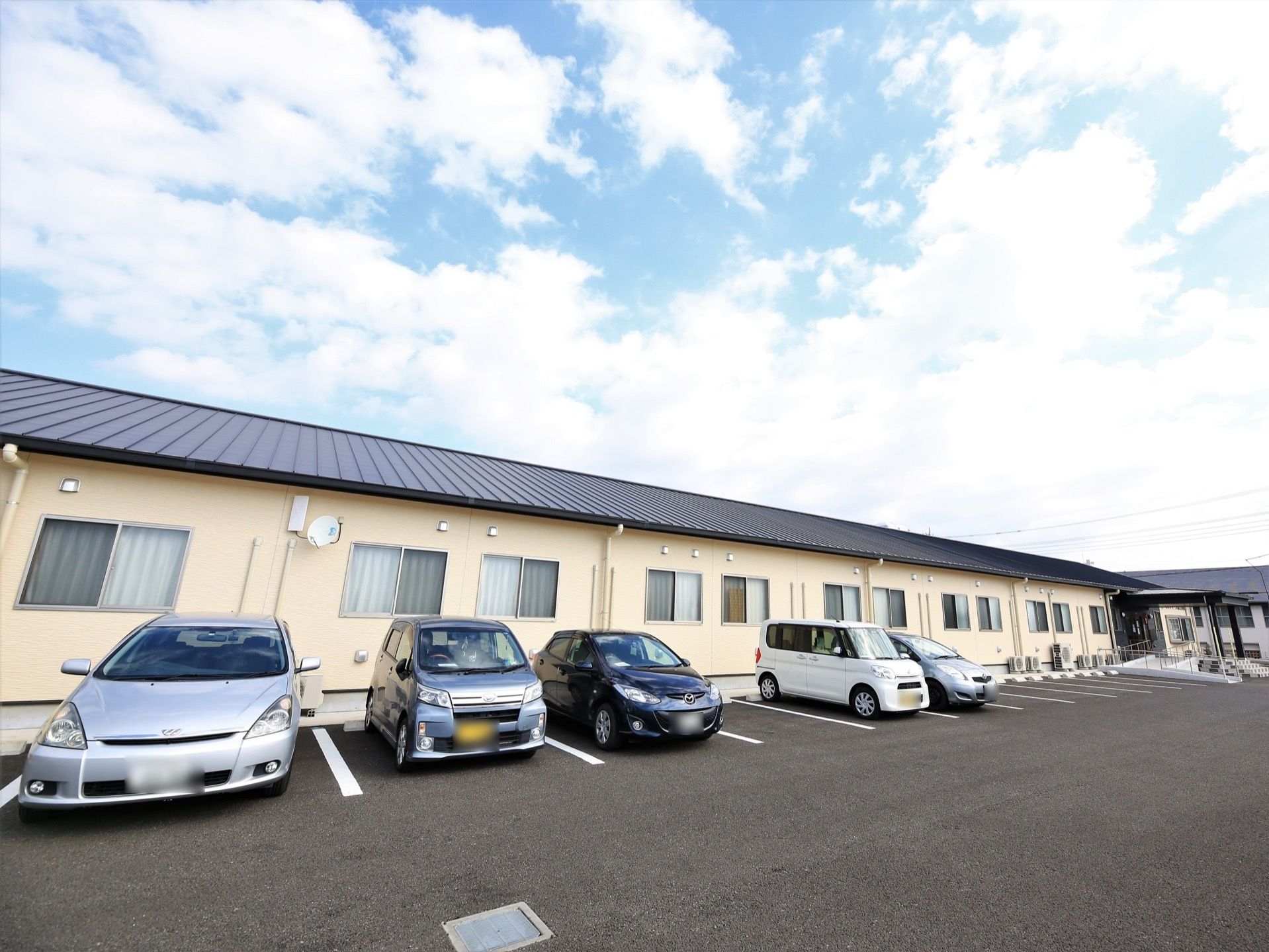 クリーム色の外壁に黒い屋根の平屋建ての建物が建っている。建物の前にアスファルトで整備された駐車場があり、数台の車が停められている。