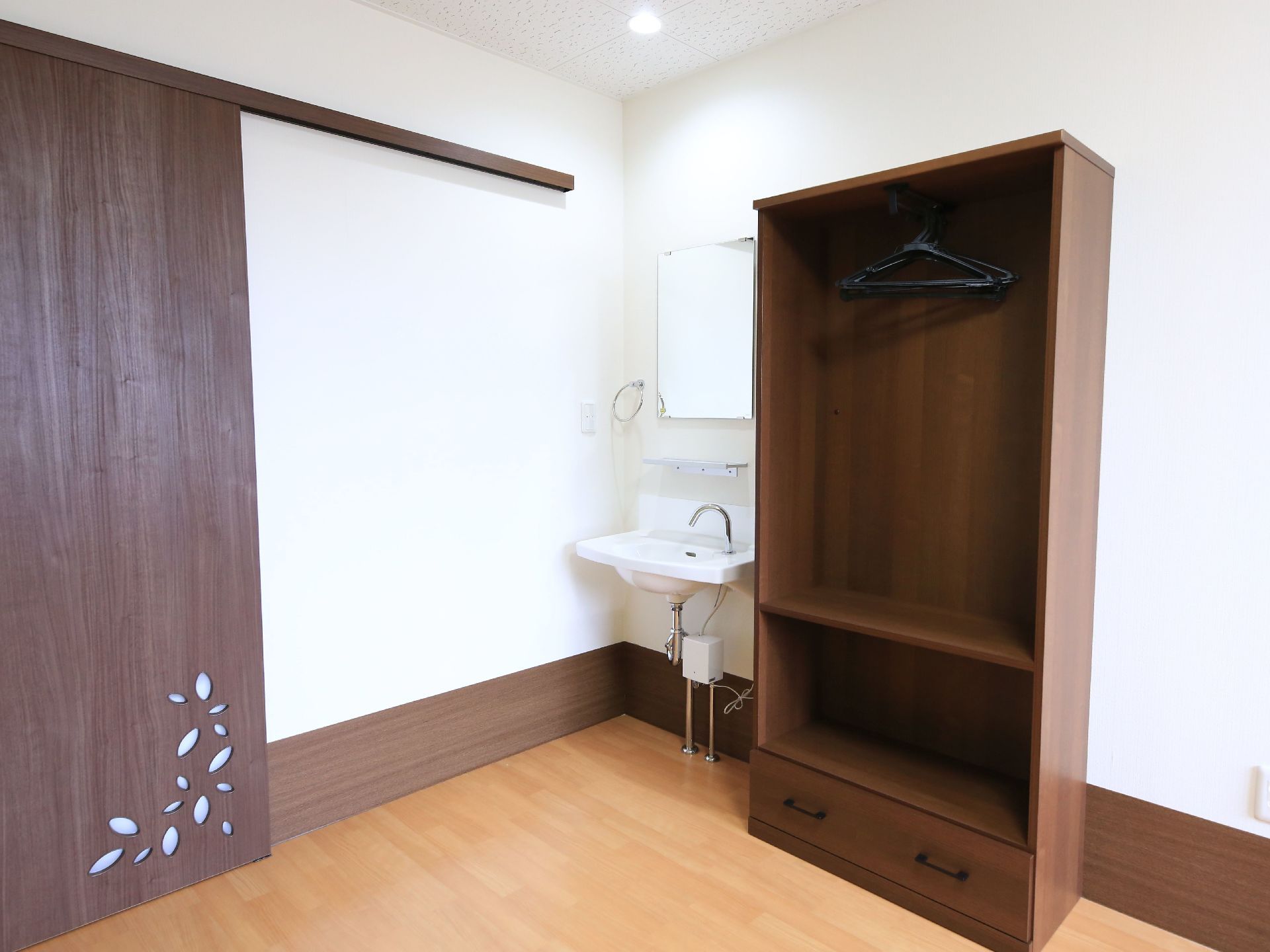 居室の角には鏡のついた洗面台が設置されている。その脇には木目仕様の大きな棚が配置されており、衣類を吊るすことができる。