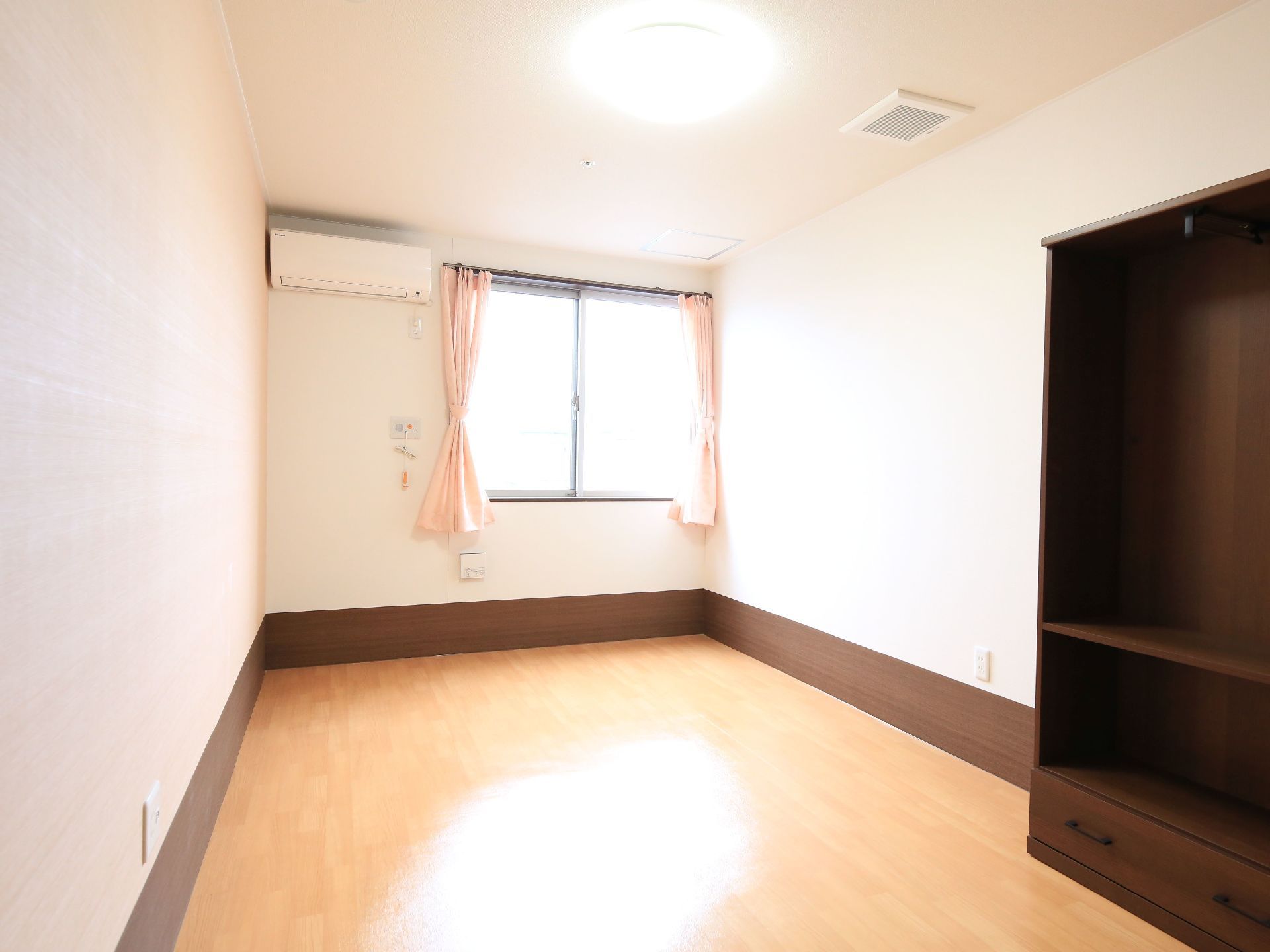 居室の明るい窓辺には柔らかいピンクのカーテンが掛かっており、日照を調節することができる。空調とナースコールが備わっている。