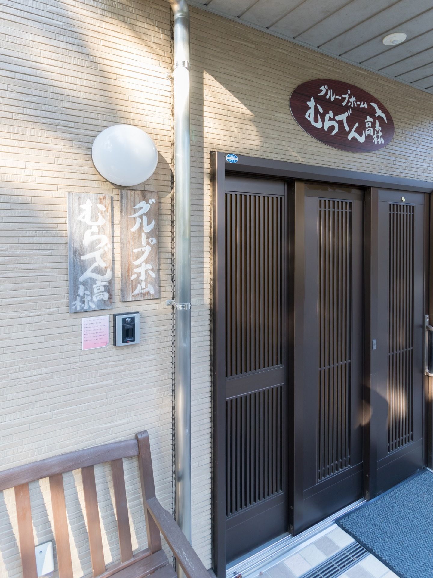 玄関出入口の上部に焦げ茶色の楕円形の表札を掲げている。横にも、縦長の板に施設種類と施設名書いて掛けている。上に照明がある。