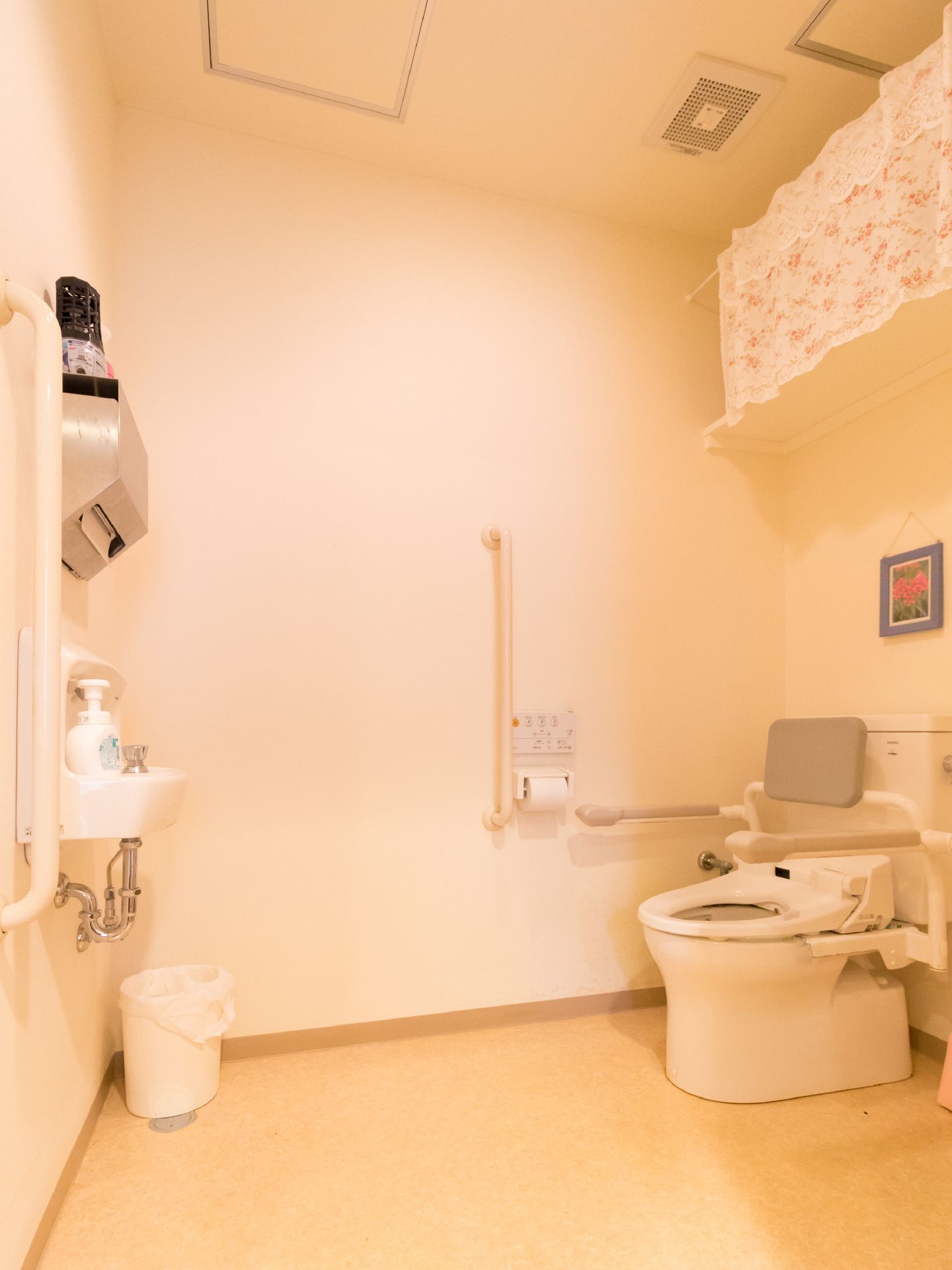 洗面台が備わった広いトイレである。温水洗浄便座や背もたれ付き肘掛け、手すりが完備されているので、快適にご利用いただける。