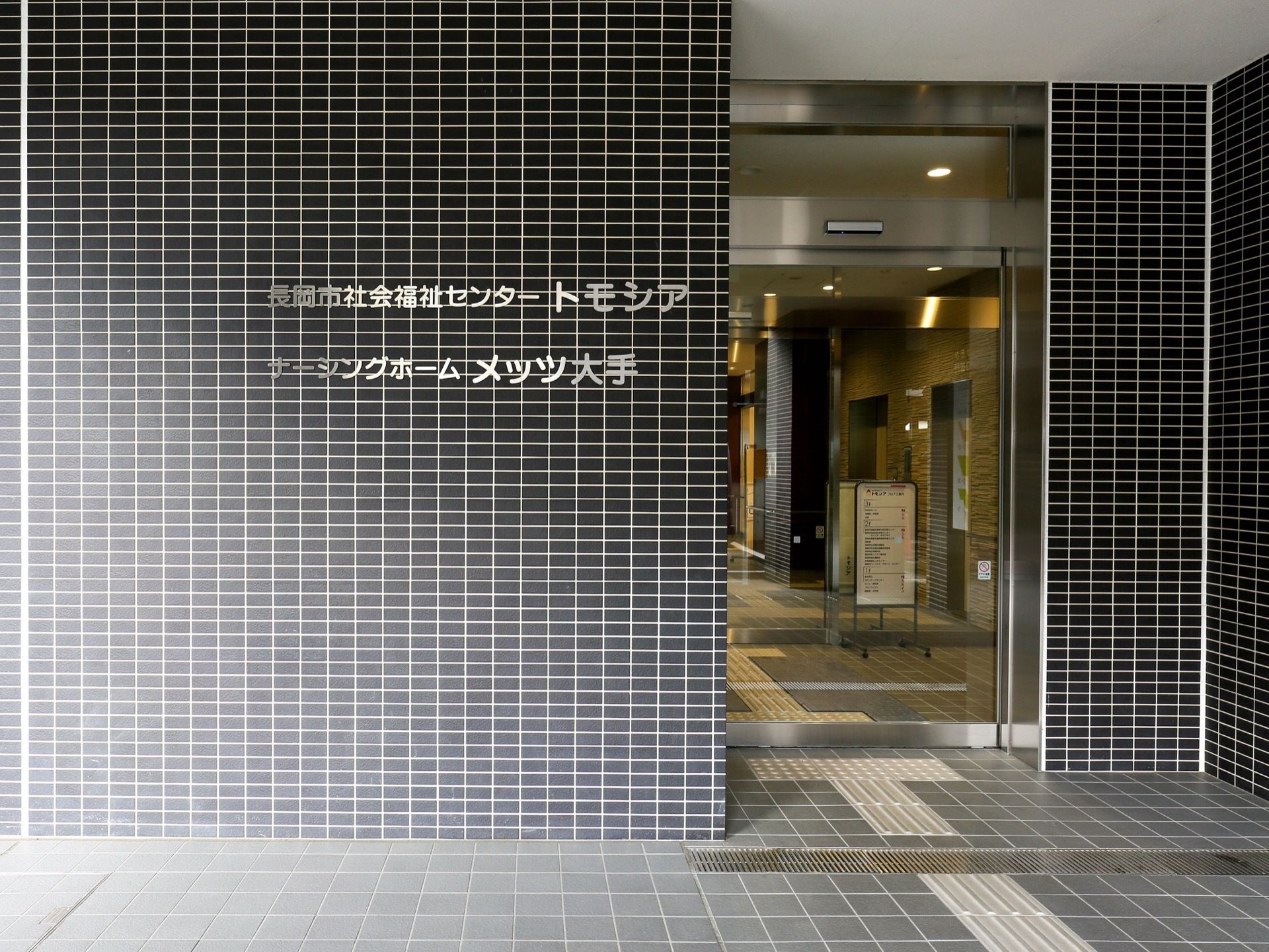 ダーク調にまとめられた、シックなエントランスである。外壁に施設名が書かれている。点字ブロックが敷かれている。
