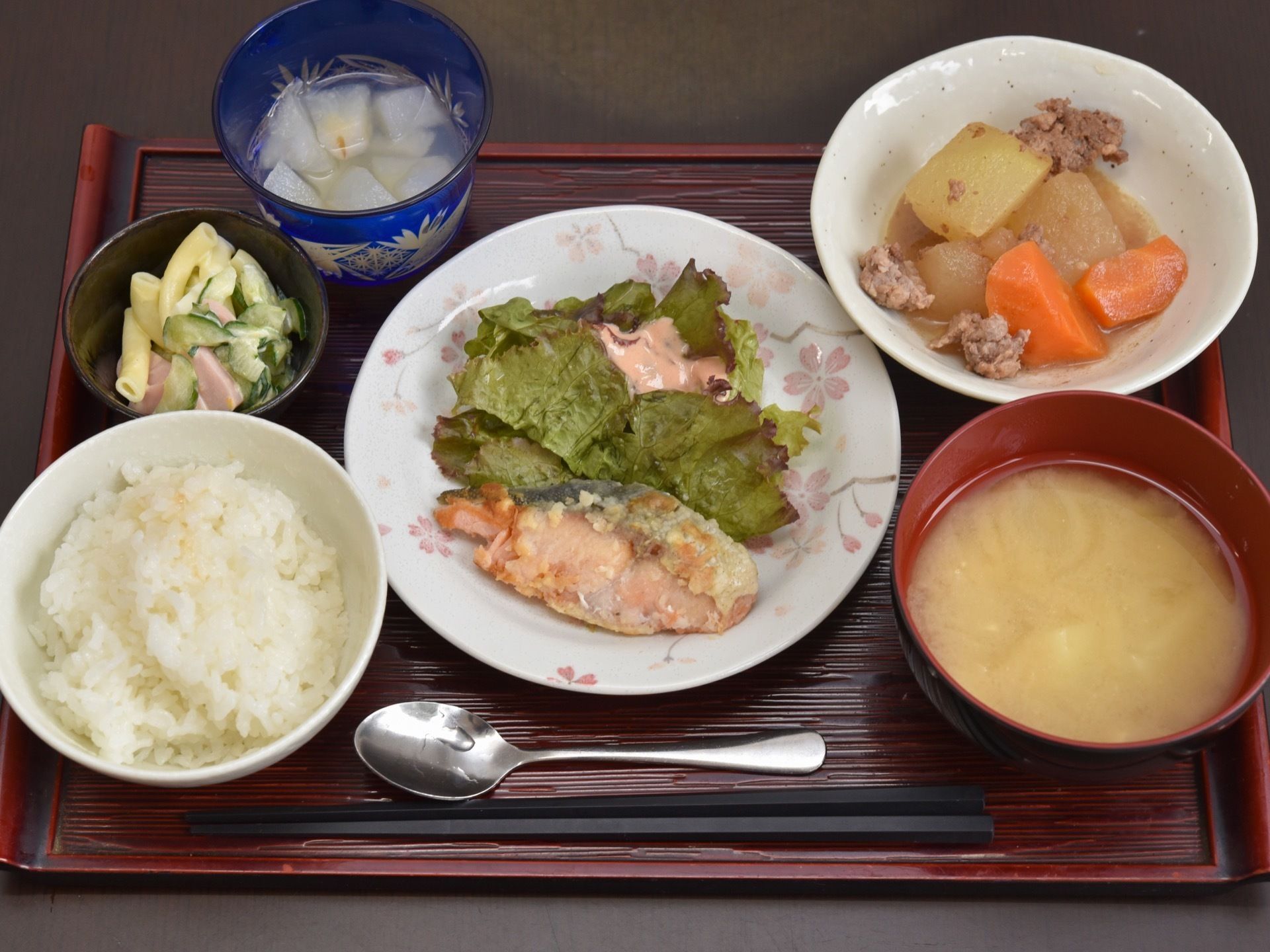 お盆に食事がセットされている。左に白いご飯、右に味噌汁、手前に箸とスプーンがある。中央にメインの料理、副菜、小鉢サラダ、デザートもある。