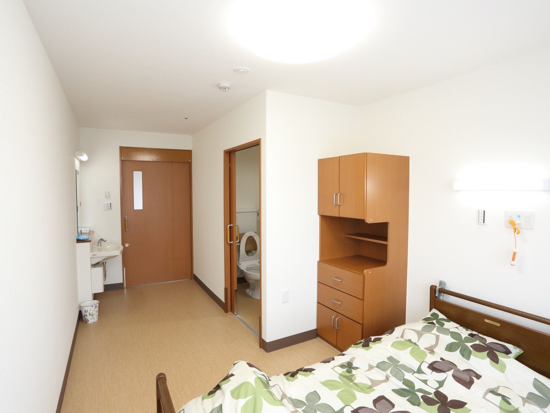 入居者様の居室にはトイレや洗面台の設備が整っており、ベッドや収納棚などの家具が置かれている。壁にはナースコールが取り付けられている。