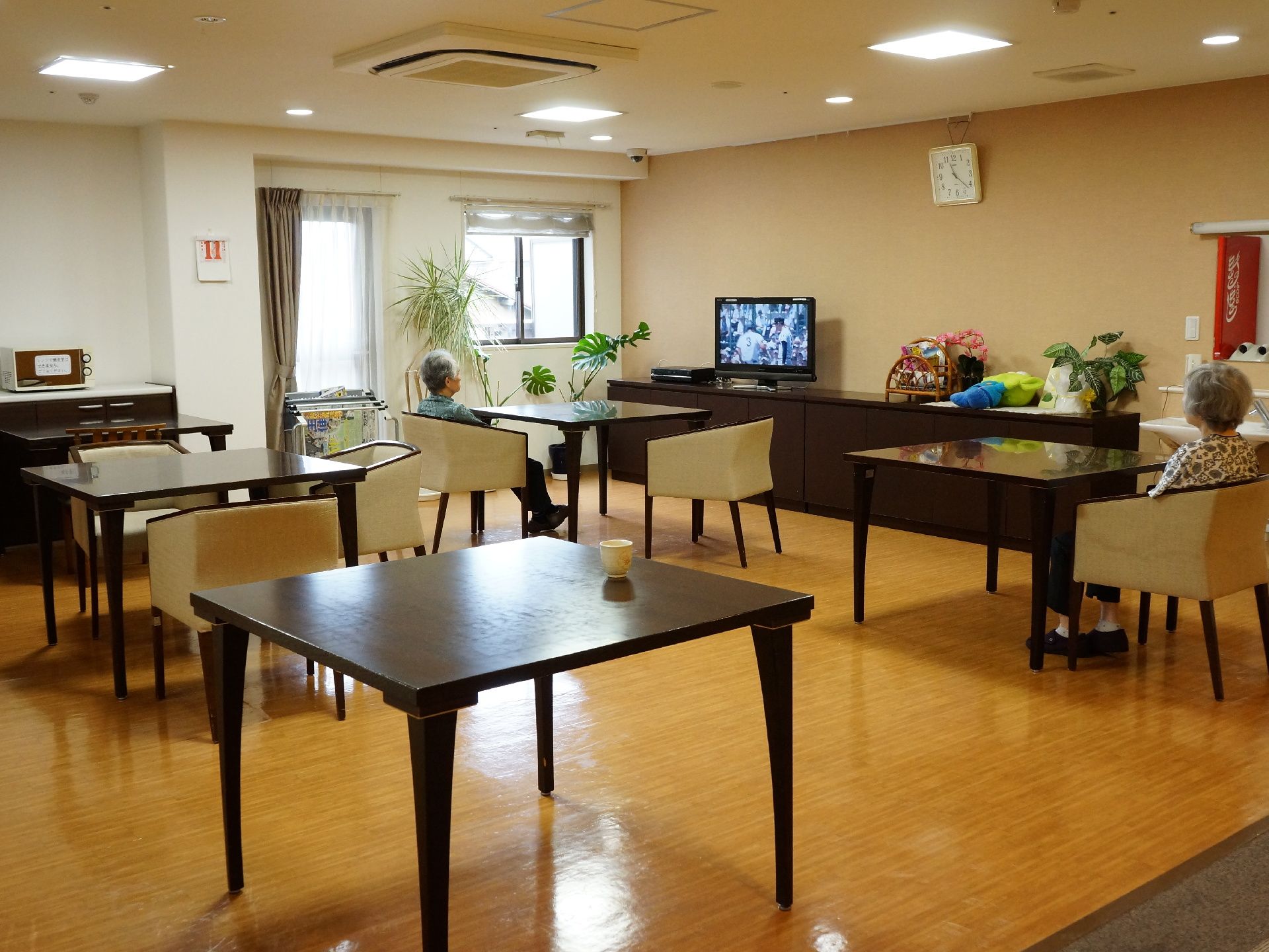 ダイニングテーブルや椅子がいくつか置かれている部屋で、壁際にテレビがある。洗面台やナースコールも設置されている。