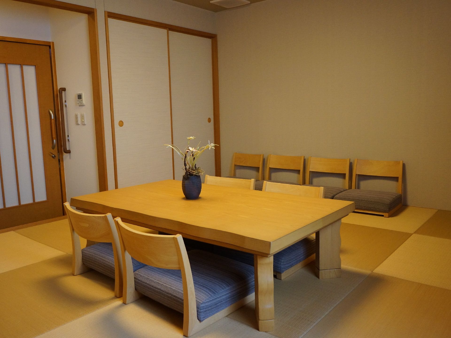 和風の引き戸のある部屋で、扉の横には押入れがある。部屋の真ん中に座卓とイスがあり、壁際にもイスが並べてある。