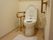 洋式トイレで、足元にはマットがひいてある。壁面と便座の横に手すりが設置されている。温水洗浄便座である。