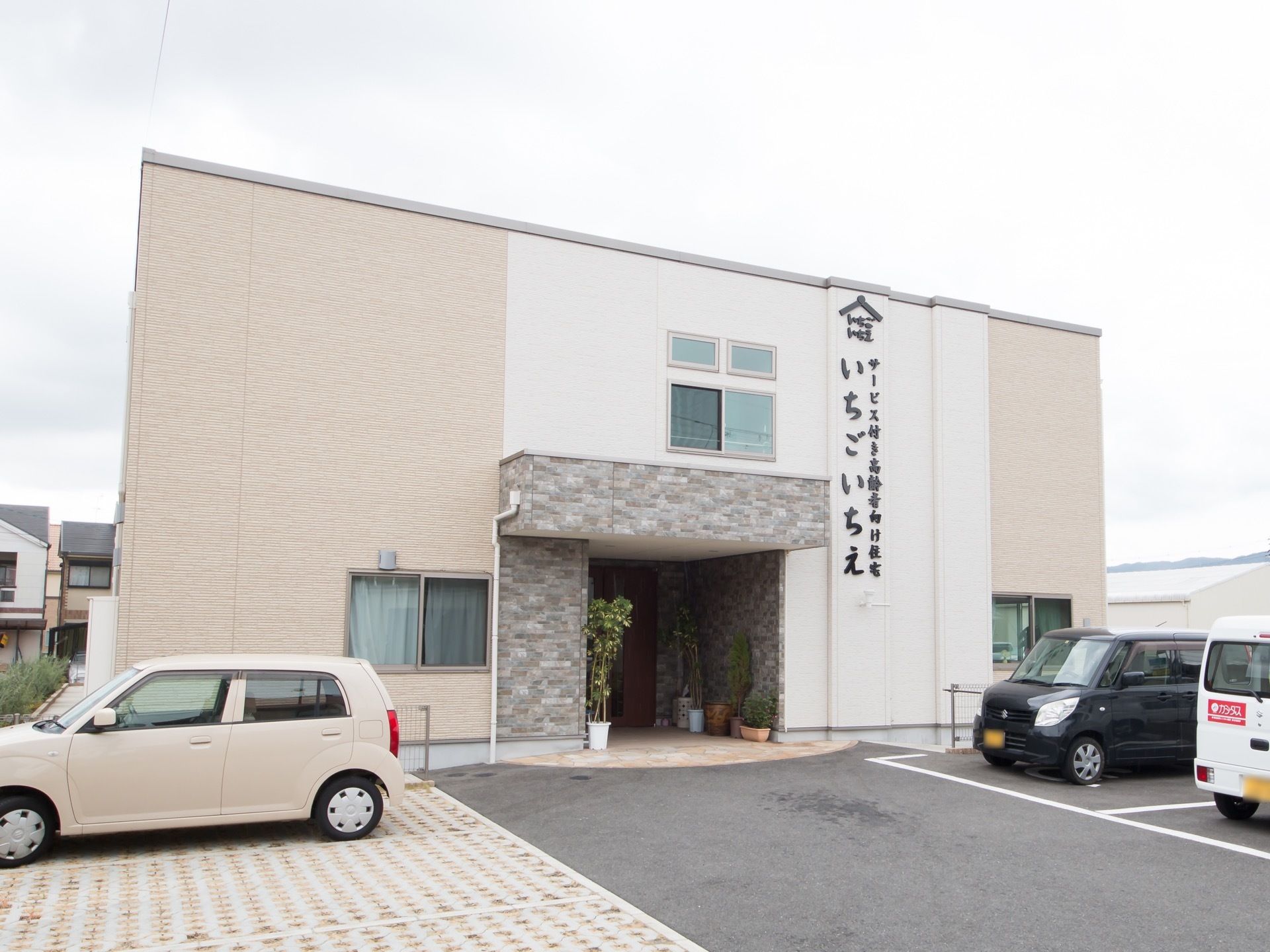 白色とベージュ色のナチュラルな雰囲気の２階建ての建物である。アスファルトとタイルで舗装された駐車場に車が停めてある。