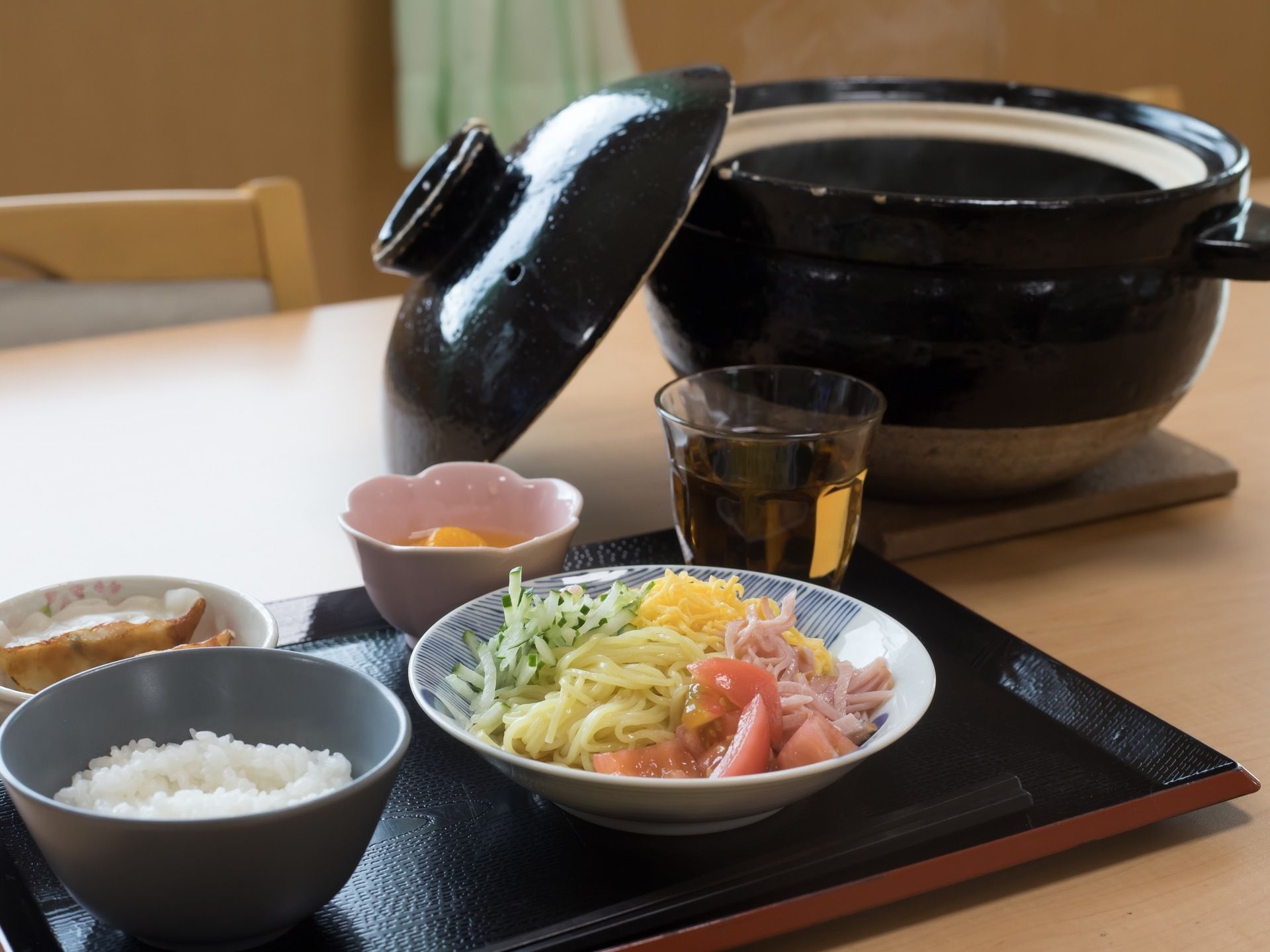 黒い配膳盆の上に冷やし中華や餃子や白米が入ったお茶碗が並べられている。黒い土鍋が置いてあり、蓋が開いている。