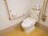 白いタンク付きの洋式便器の上に予備のトイレットペーパーが重ねて置いてある。壁に可動式の手すりと固定の手すりが取りつけられている。
