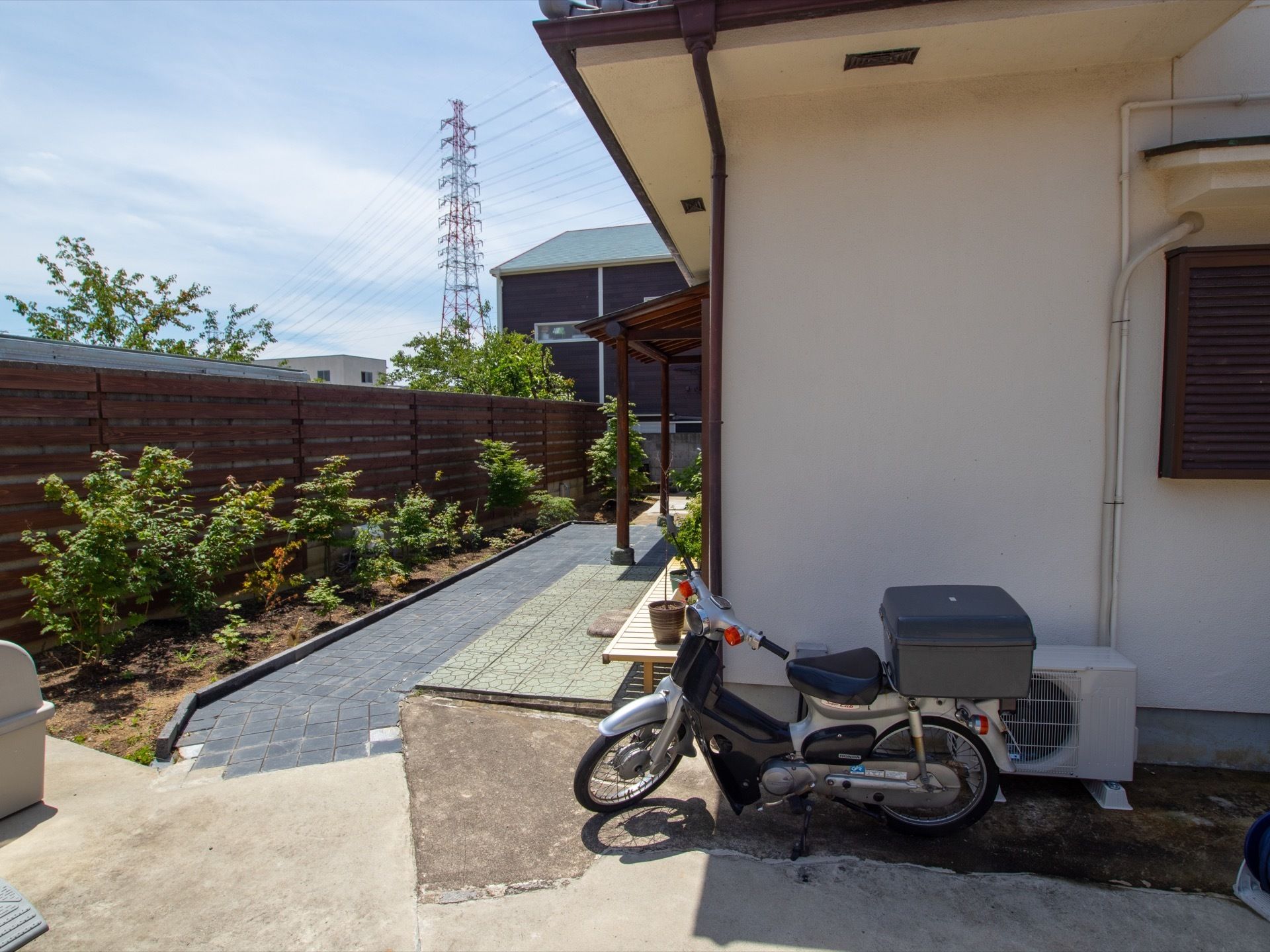 周辺には高い建物がない。縁側の近くにバイクが停められている。木製のついたての前には多くの木々が植えられている。