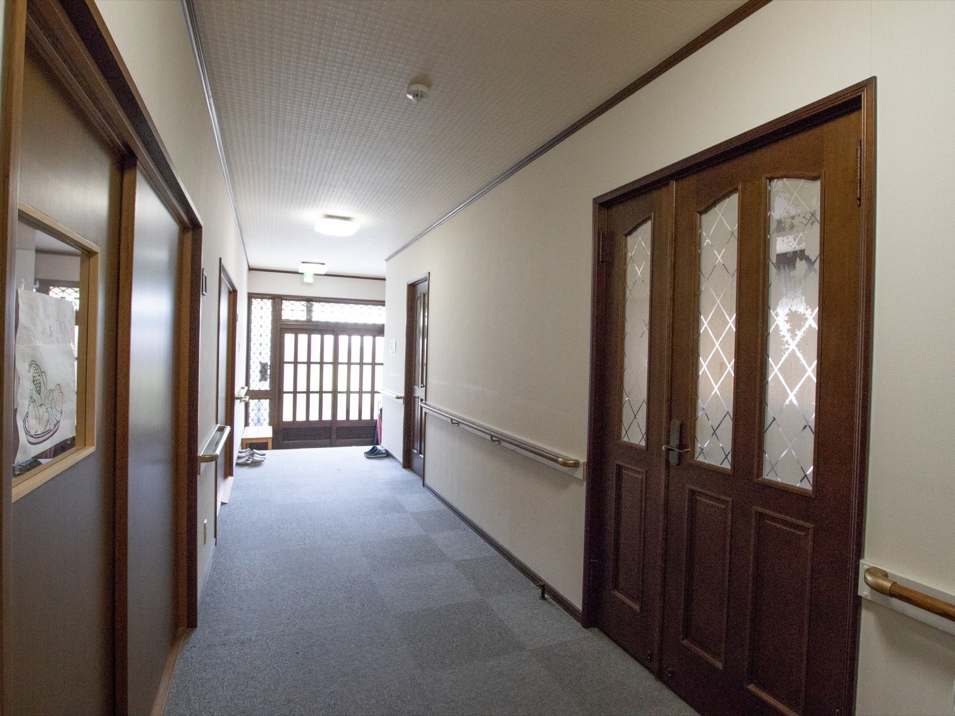 廊下は横幅が広い。壁には手すりが設置され、足元には格子状のグレーの絨毯が敷きつめられている。玄関からは外光が差し込んでいる。
