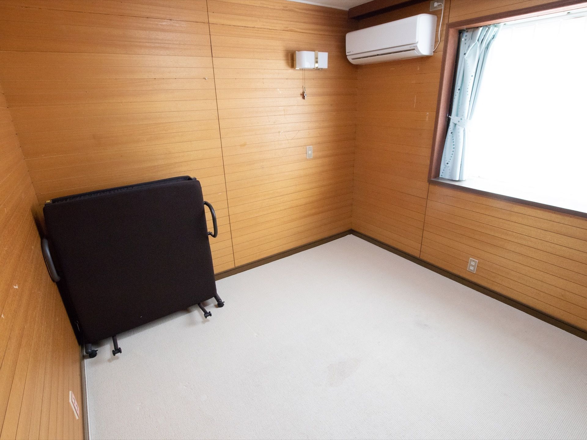 茶色の壁にエアコンと大きな窓の個室がある。窓には水色のカーテンがかかっている。壁際には黒の折り畳み式ベッドが立てかけてある。