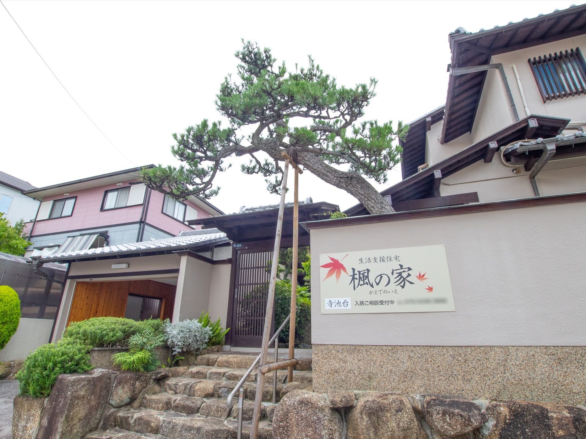 自然石を使った雅な石を使用した階段がある。二階建ての日本家屋と、ひさしの付いた塀が建物を囲っている。