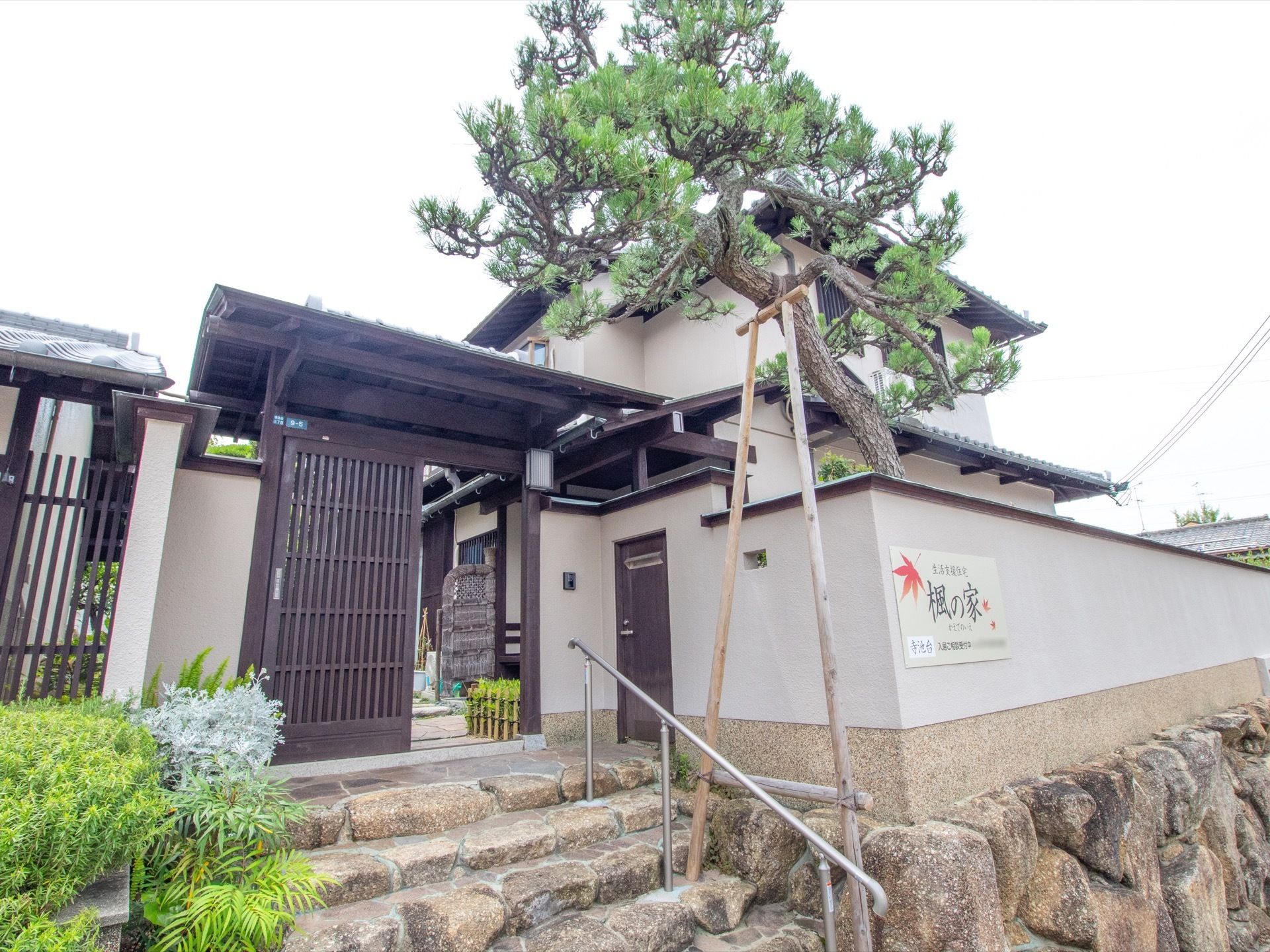 石段と日本風の塀と石垣に囲まれた二階建ての日本家屋がある。入口は格子戸になっており、横には松の木がある。石段には手すりが設置されている。