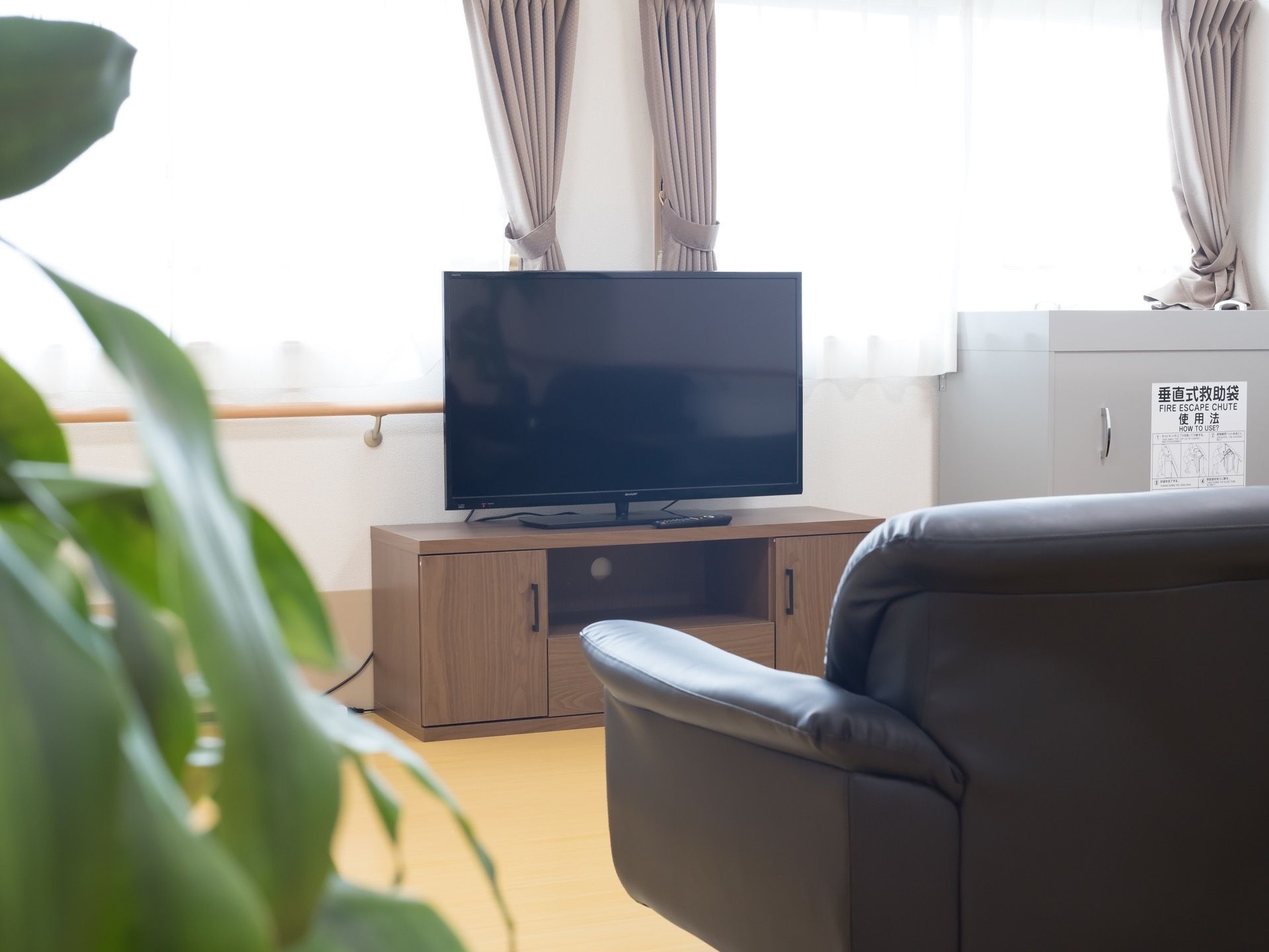 黒いソファのが置かれ、前には木目調の台の上にテレビが設置されている。窓にはレースカーテンが引かれ、その下には手すりが取りつけられている。