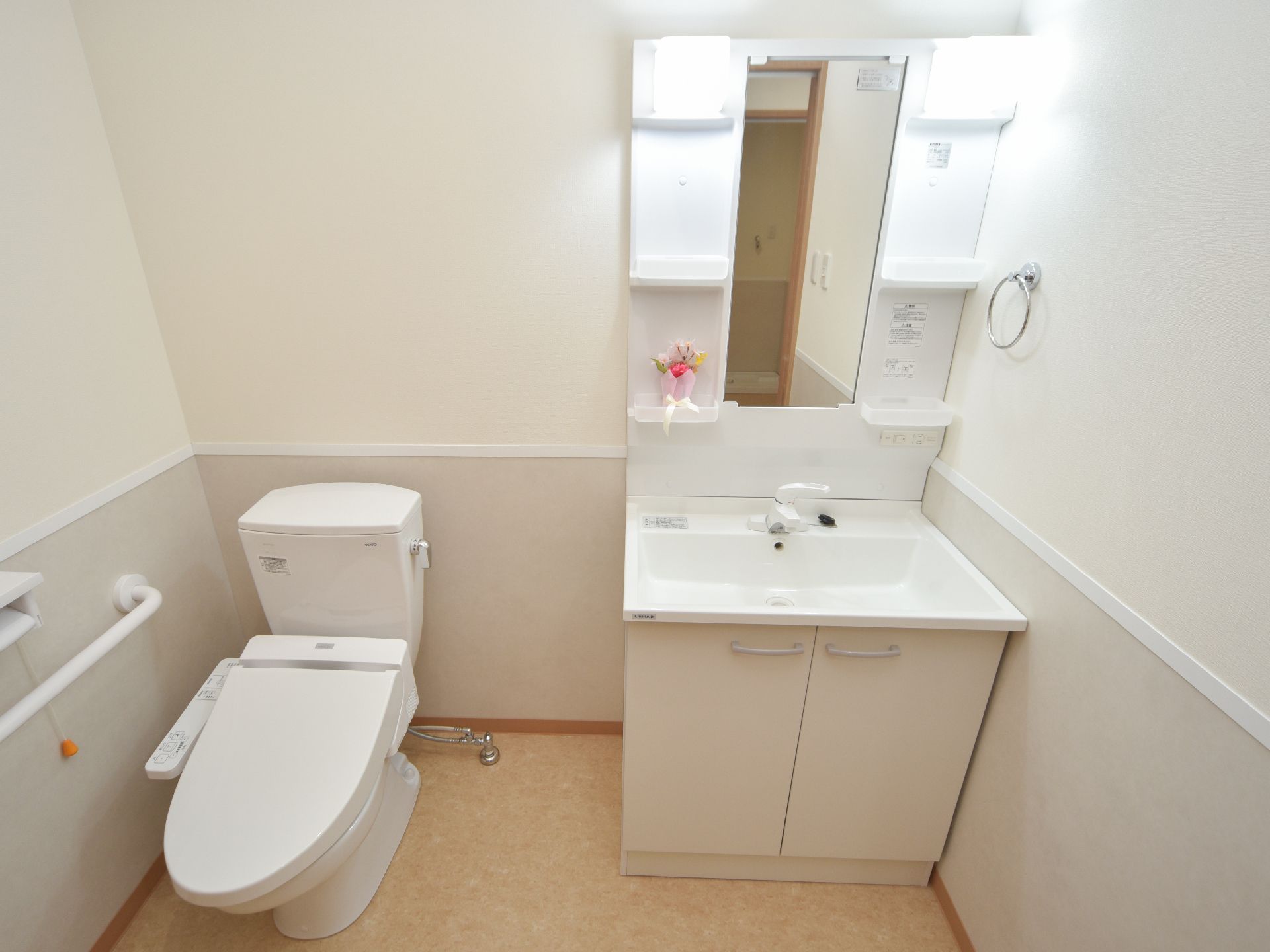 トイレスペースの便器から少し離れた横に、洗面器、収納、鏡が一体になった家庭向きの洗面台がある。洗面台の右壁にタオルホルダーがある。