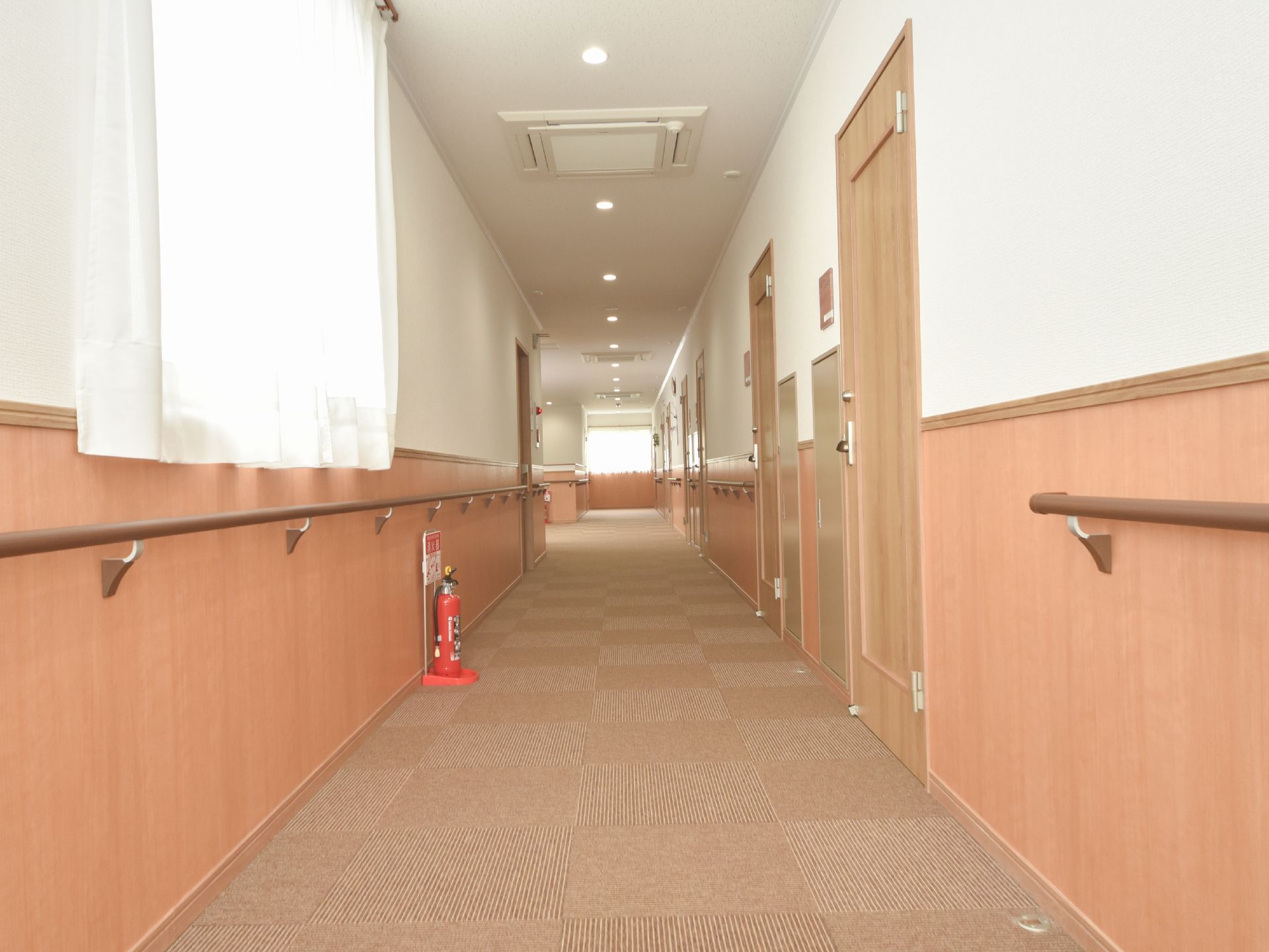 廊下の床、部屋の扉、壁の腰高の位置までベージュ系にまとめられている。壁には手すりが続いている。所々に腰高窓がある。