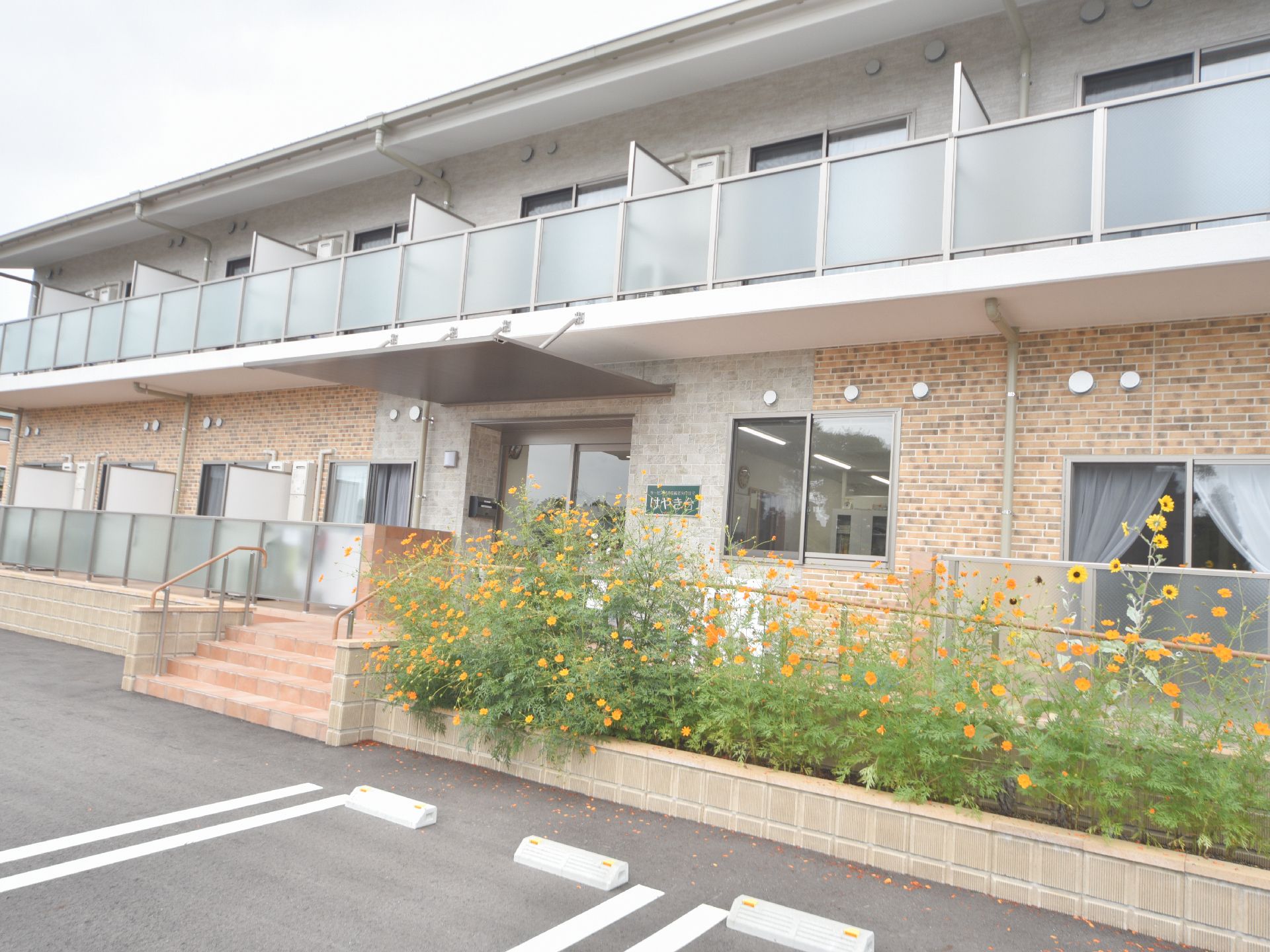 2F建ての施設の壁面は、レンガ調のオレンジ系と白系のツートンの組み合わせ。全部屋ベランダが付いている。建物を囲む塀は花壇もある。
