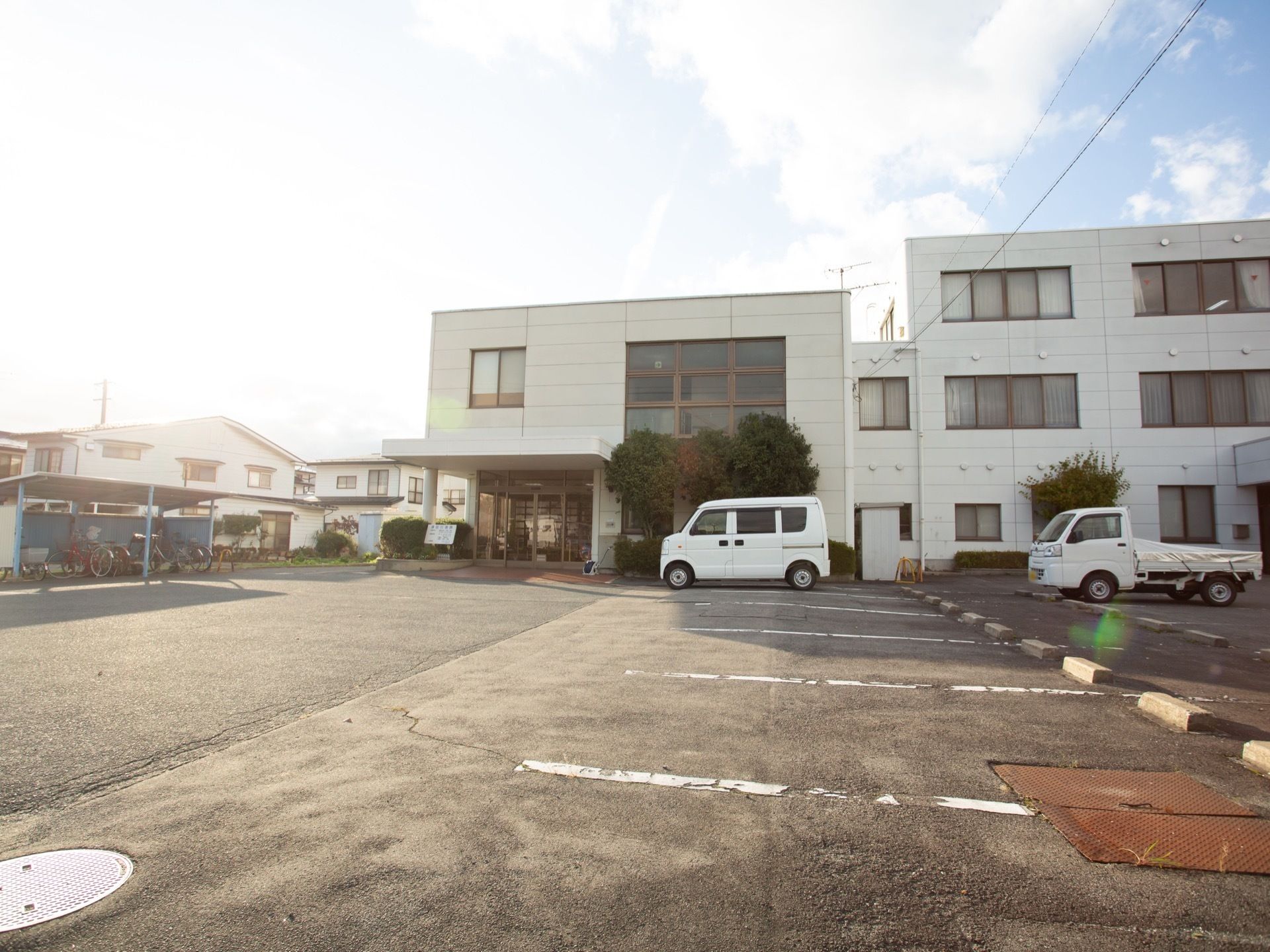 白色の壁の３階建ての建物で、敷地内に広い駐車場が整備されている。駐車場の脇に、駐輪スペースも備わっている。