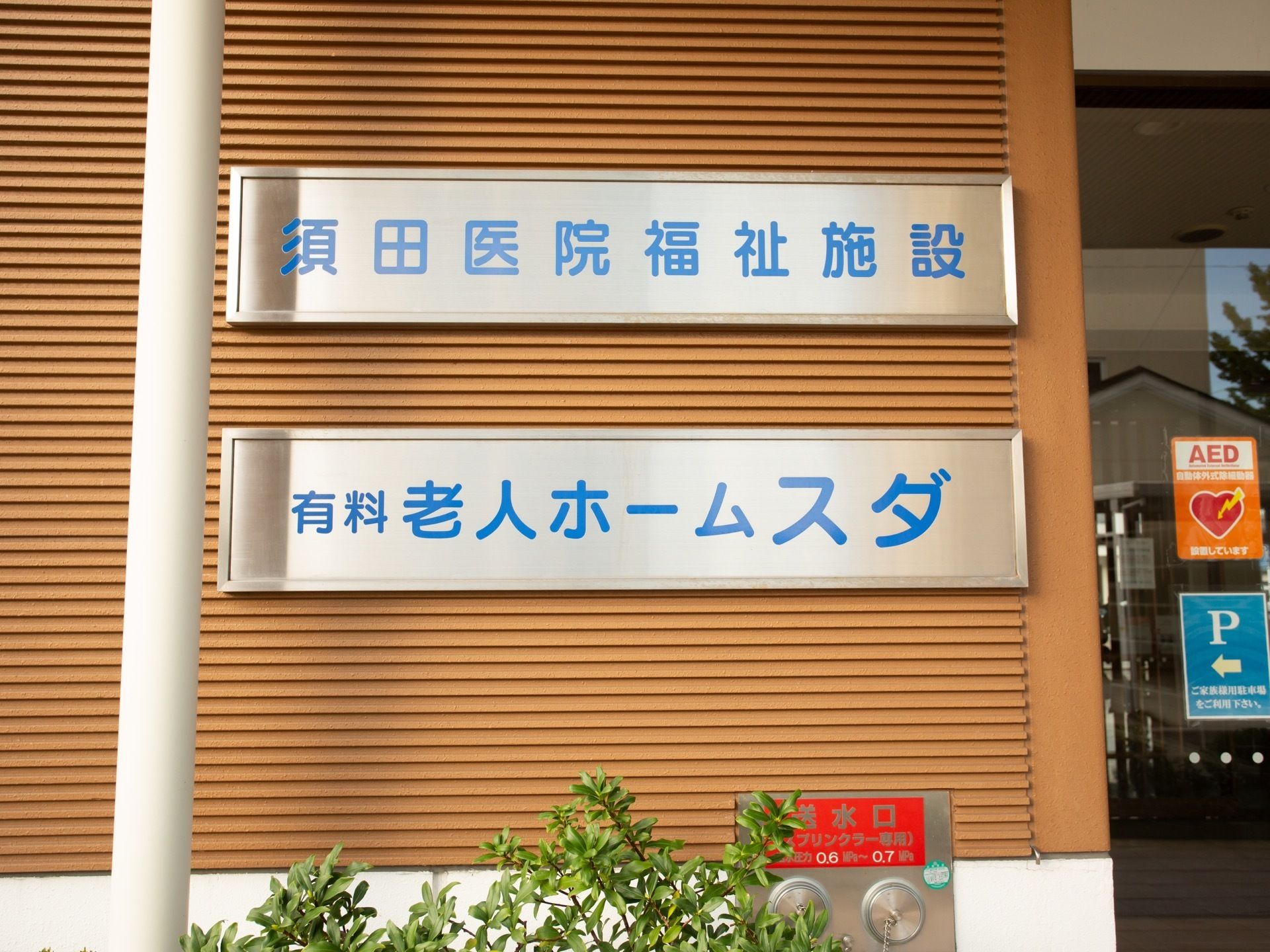 玄関の入り口の横に、横に長いシルバーのプレートに青色の文字で施設名が書かれた看板が二枚掲げられている。