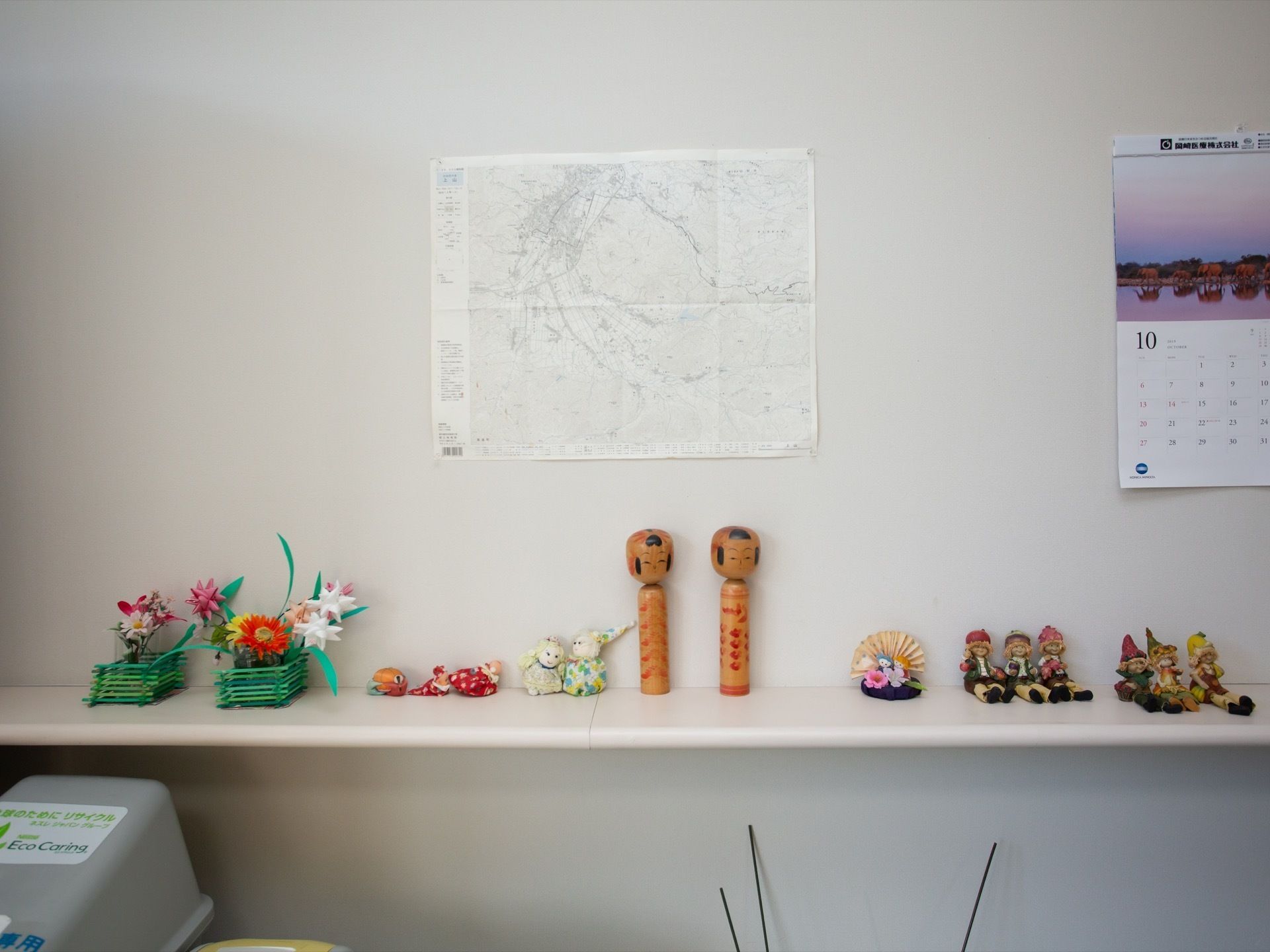 飾り棚の上に、こけしや小さな人形、花の飾りなどが並べられている。壁には地図やカレンダーが掛かっている。