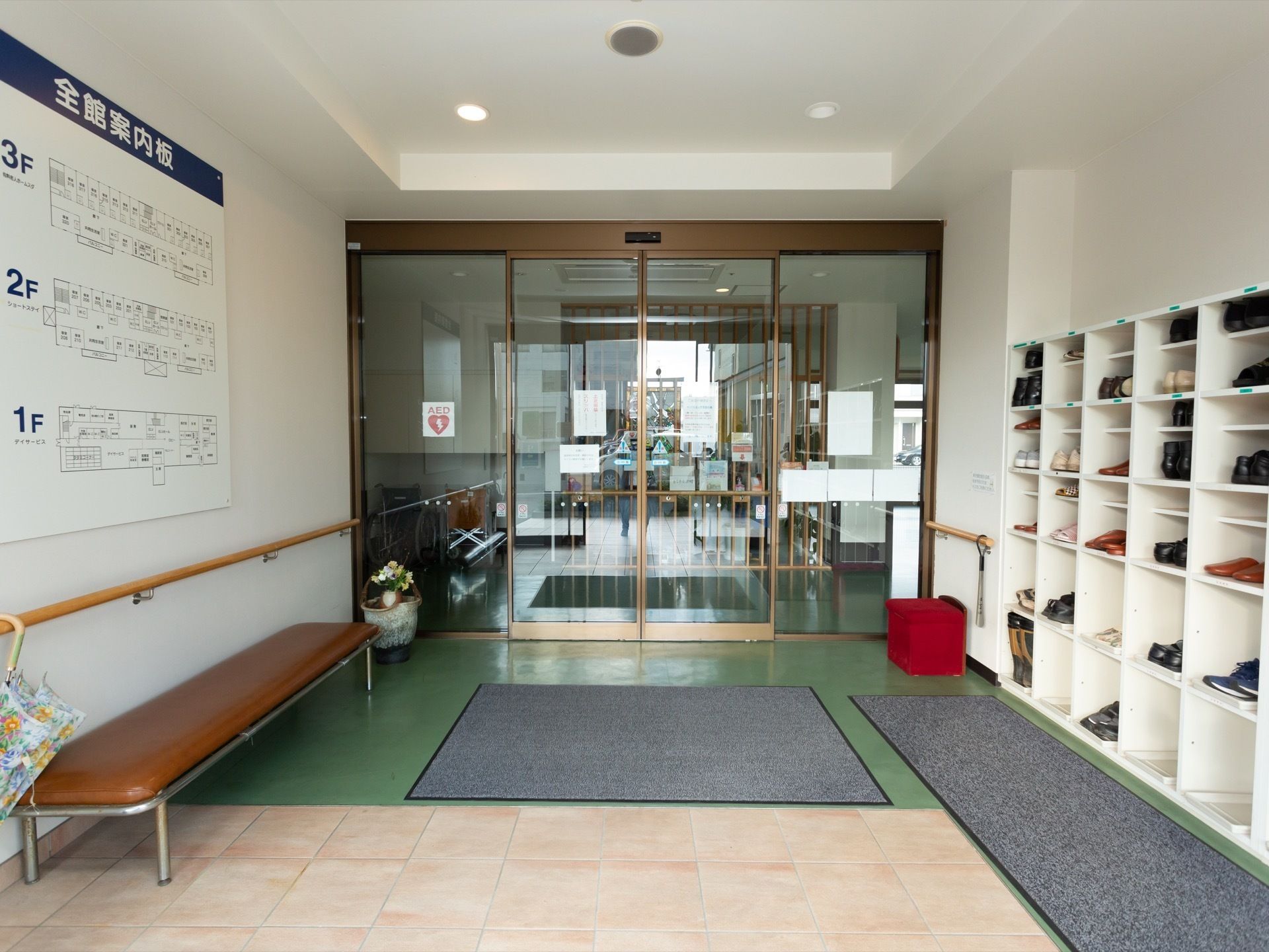 右側の壁沿いに、白色の靴箱が設置されている。左側の壁には館内の案内板や手すりがあり、ベンチが置かれている。