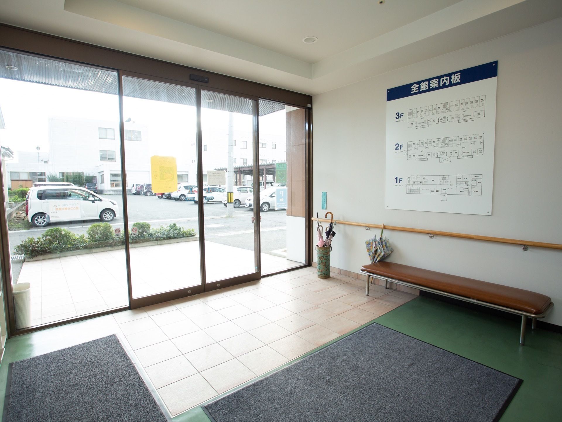 外から玄関内まで、床に段差がない。壁際に、茶色の座面の横に長いベンチが置かれている。壁に館内の案内ボードが備わっている。