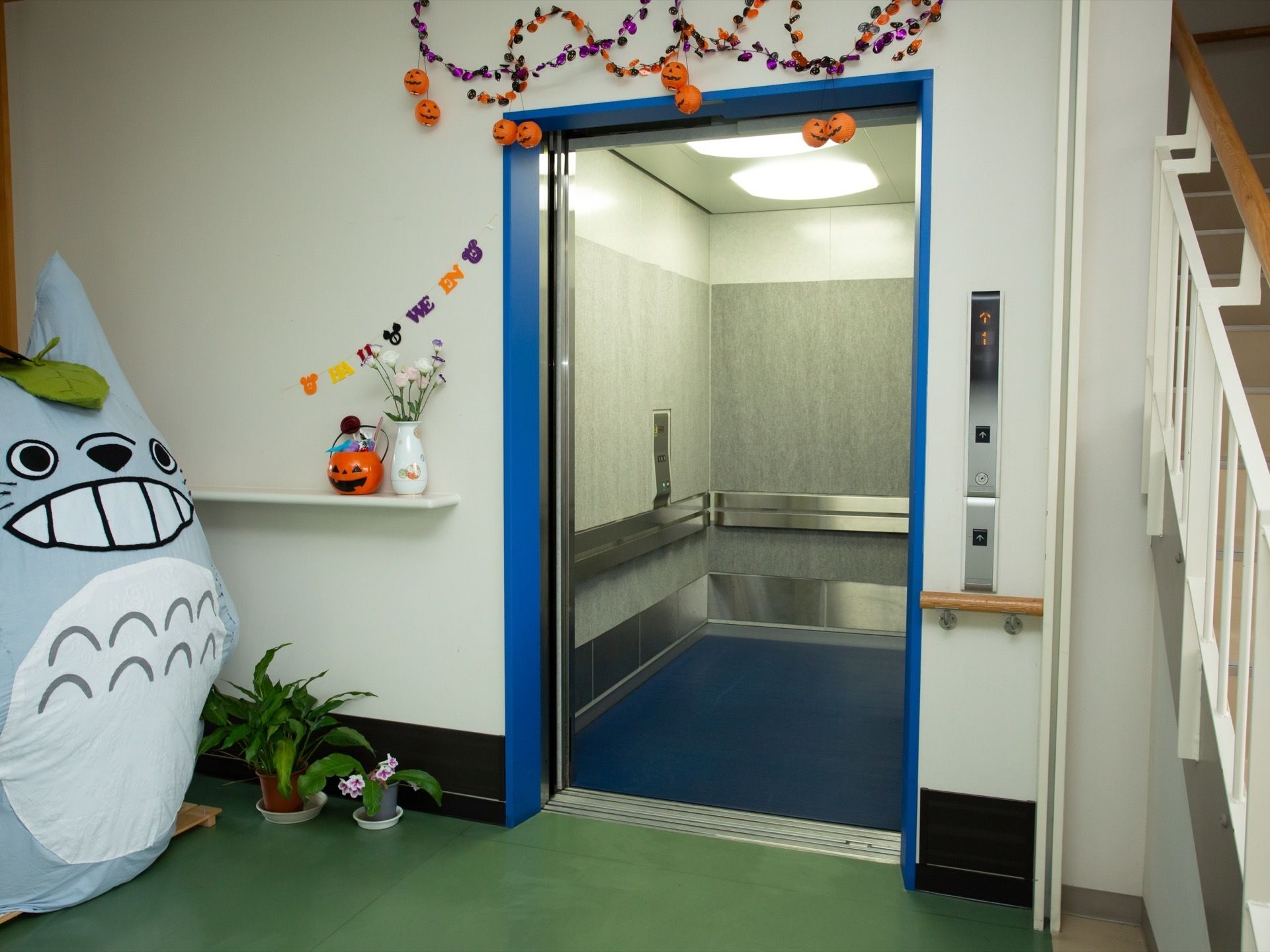 青色の縁取りのエレベーターがあり、扉が開いている。手前にはハロウィンの飾りや手作りのトトロの人形が置かれている。