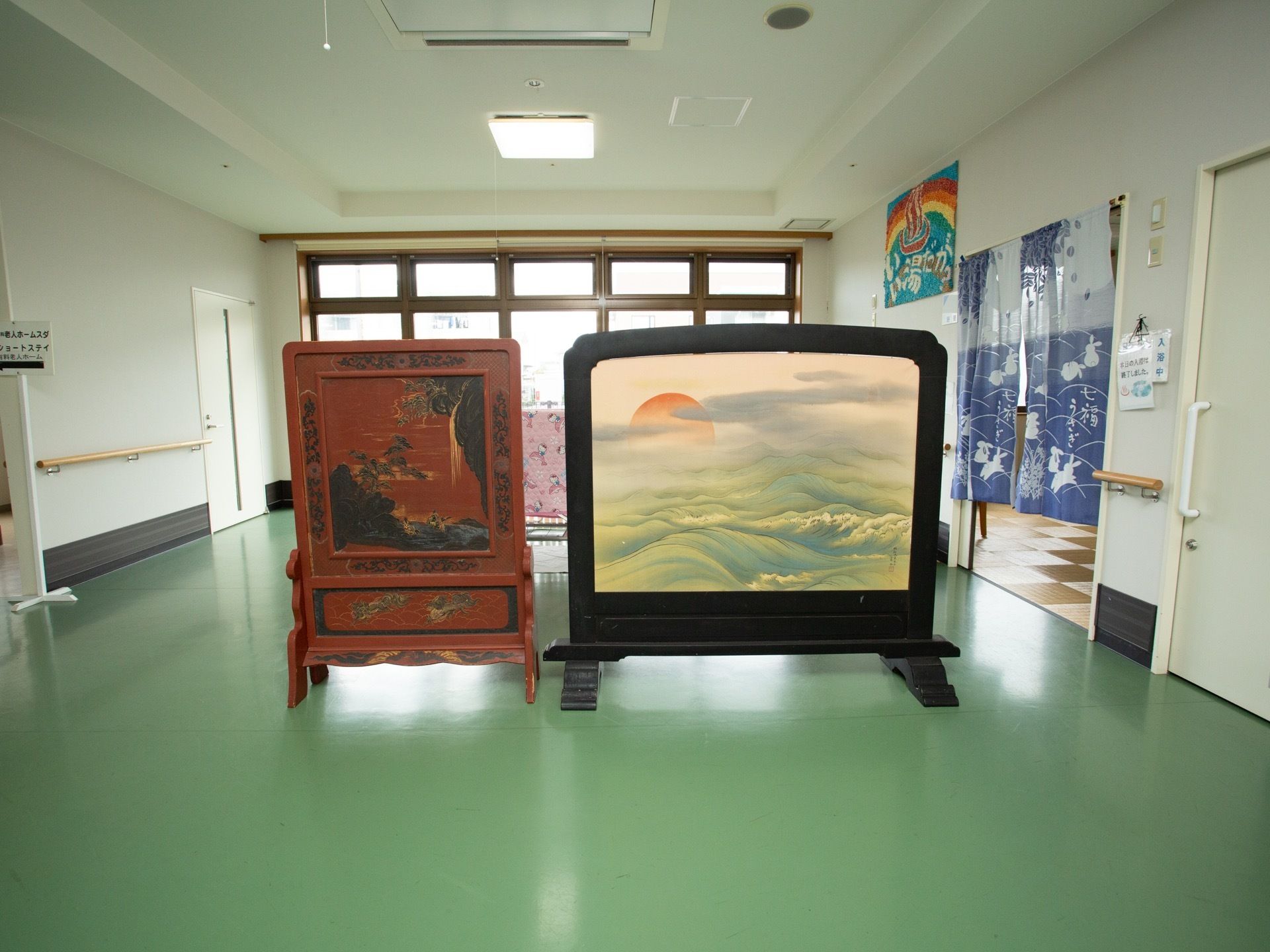 廊下の一角に、山や松がデザインされた衝立てが置かれている。淡いグリーンのビニル床で、壁には手すりがある。