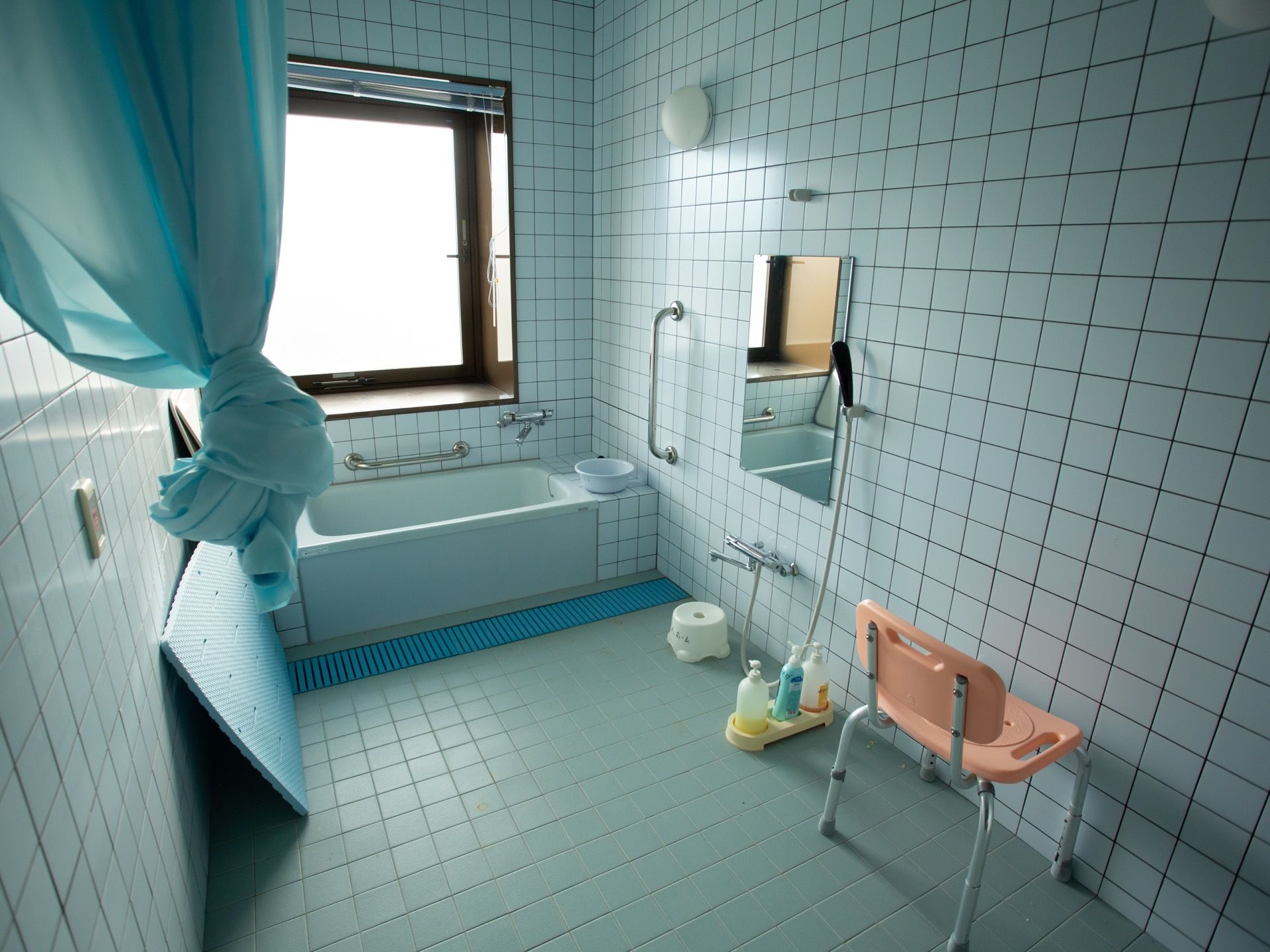 薄いブルーグレーのタイル貼りの浴室で、浴槽周りに手すりが備わっている。洗い場にシャワーチェアが置かれている。