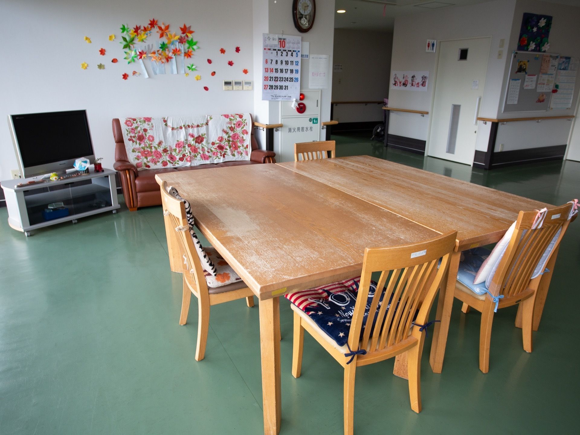 中央にテーブルが２台並んでおり、椅子が４脚置かれている。壁際にソファやテレビがあり、近くにトイレがあるのが見える。